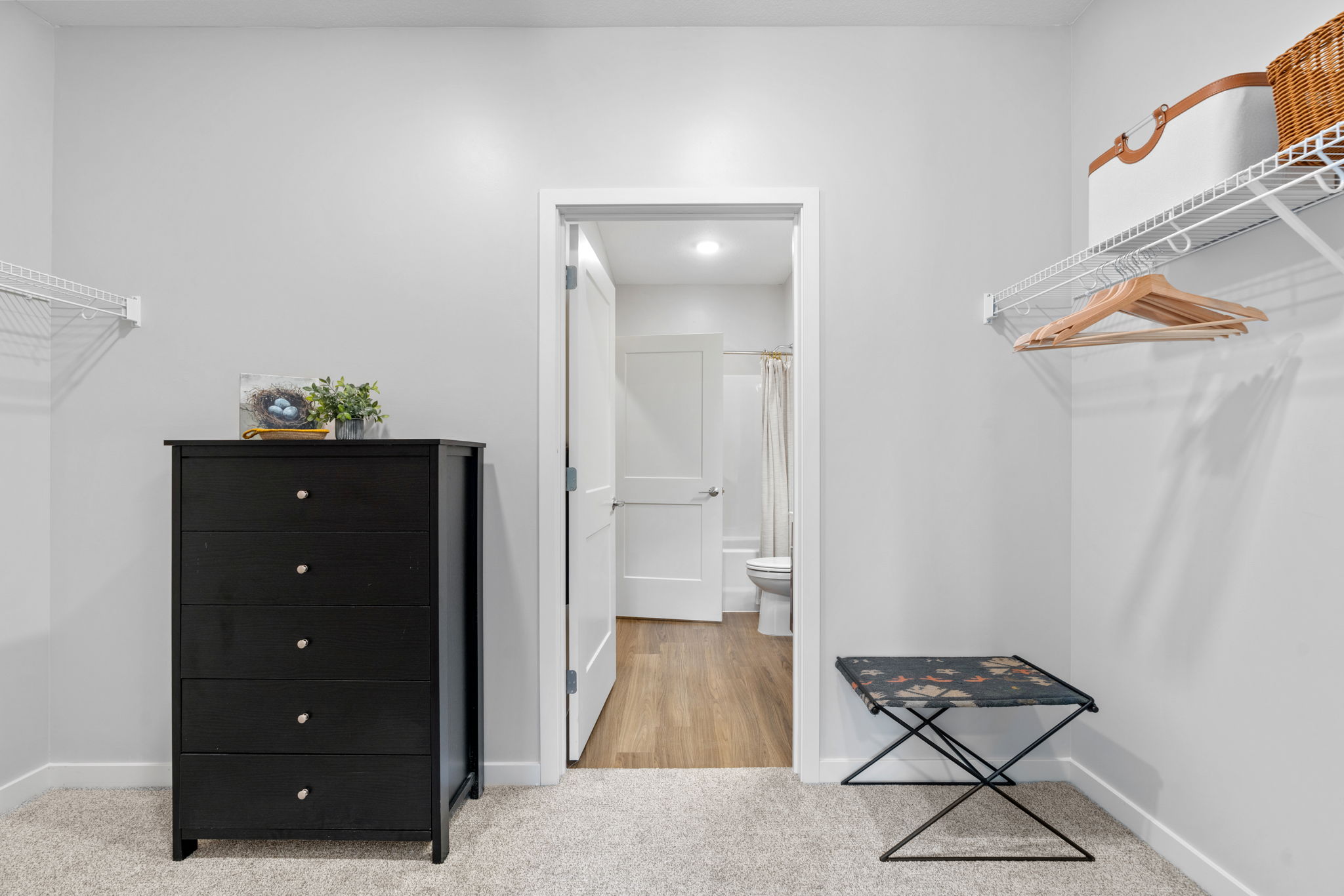 A bright, minimalist closet featuring a black dresser with five drawers, a folding stool, and wire shelving with wooden hangers. A doorway leads to a bathroom in the background, with light-colored walls and warm wood flooring. The space is neat and organized, enhancing its modern aesthetic.