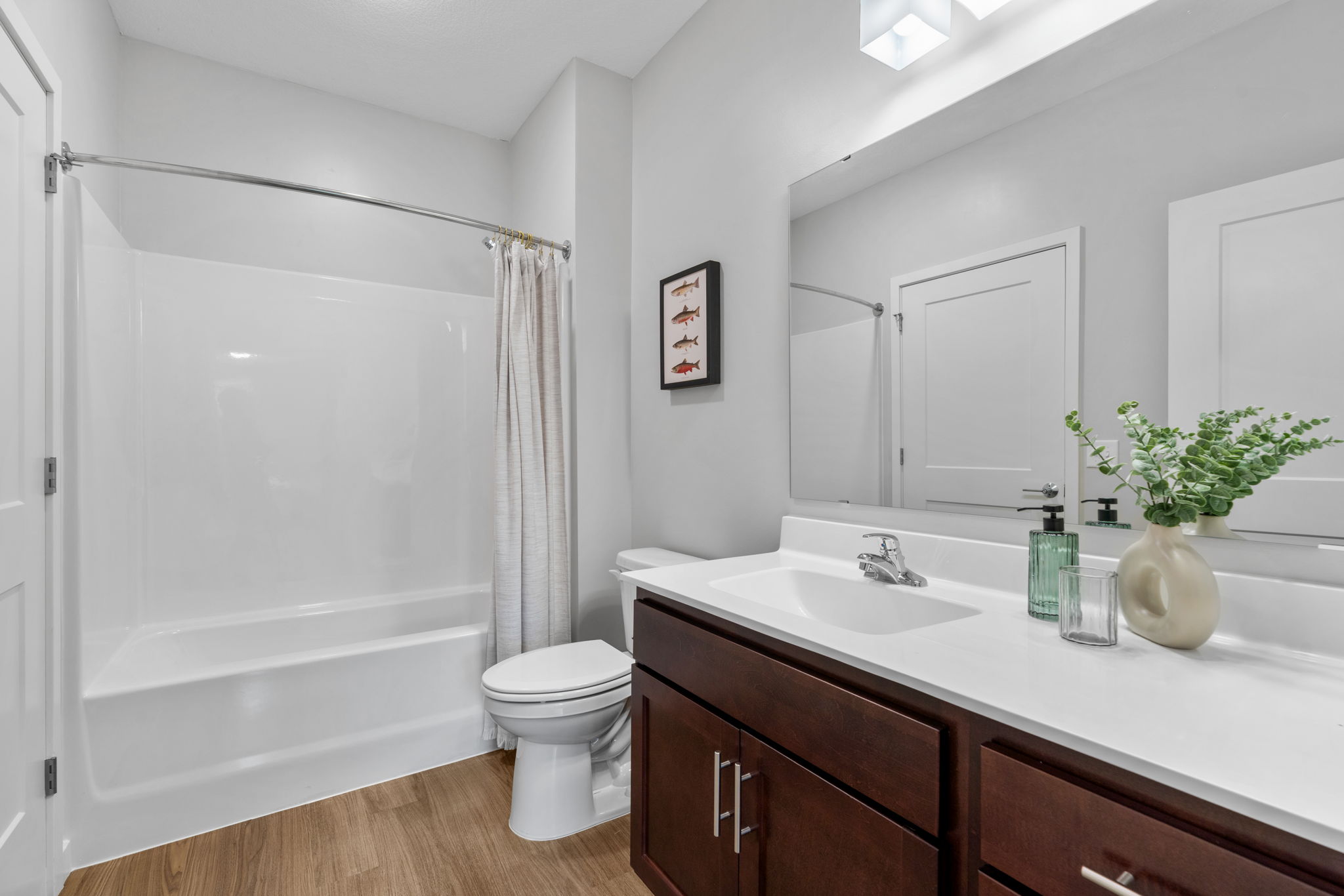 A modern bathroom featuring a bathtub with a shower, a white vanity with a sink, and dark wooden cabinets. There is a decorative plant on the counter and a framed picture on the wall. The flooring is light wood, and the walls are painted in a soft gray tone. Natural light illuminates the space.