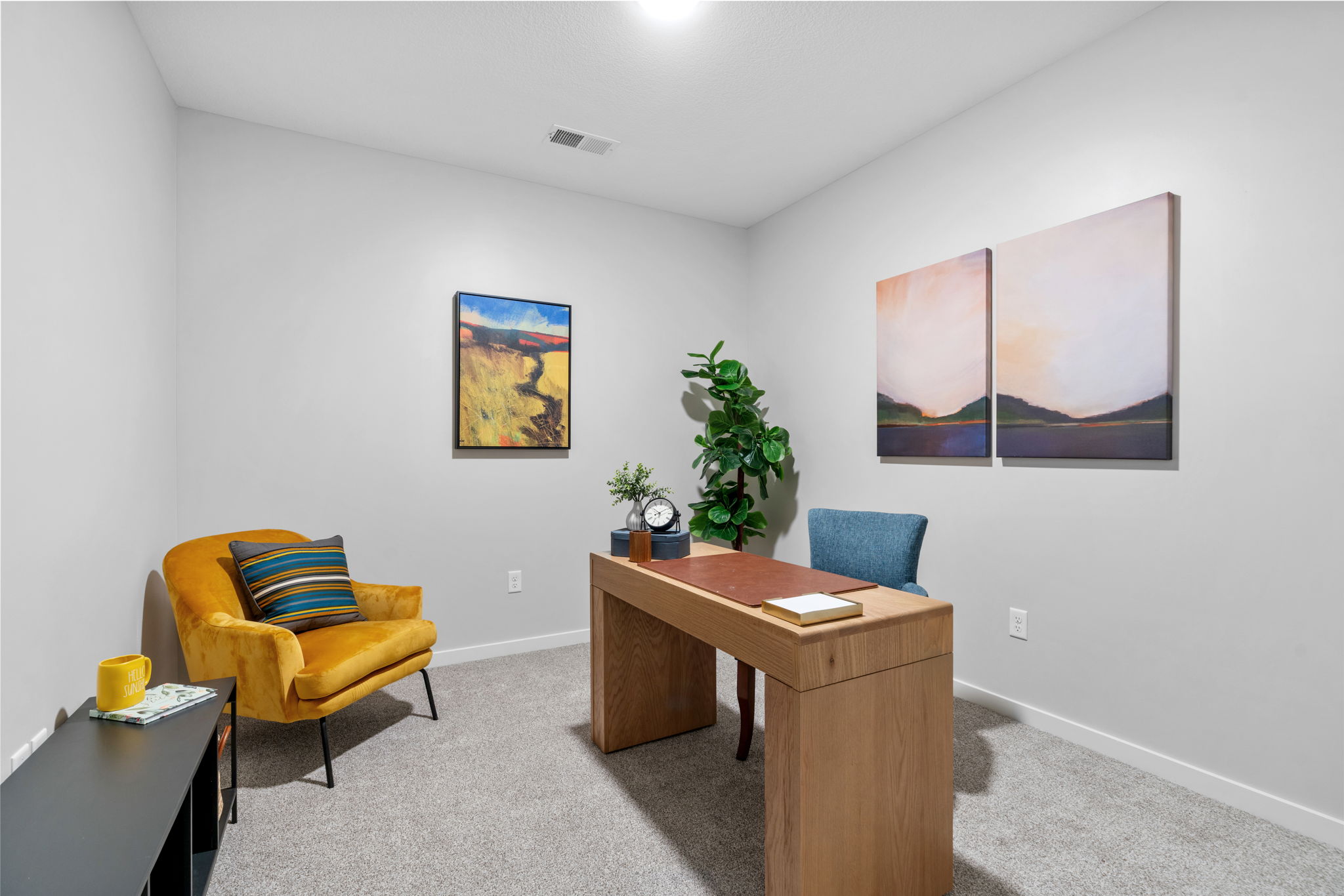 A cozy office space featuring a warm yellow armchair with striped cushions, a modern wooden desk with a small clock and a plant, and two abstract paintings on the wall. The room has a neutral color palette, carpeted flooring, and ample natural light from an overhead fixture.