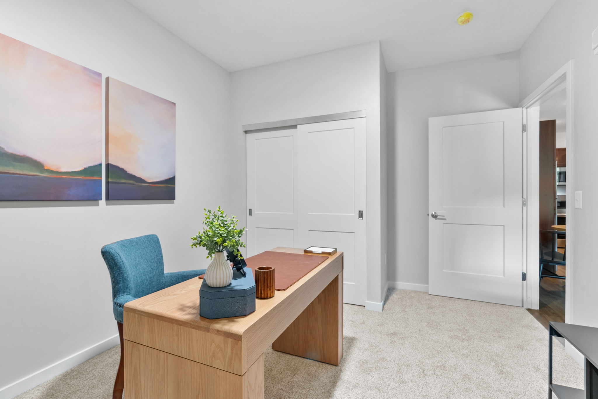 A modern office space featuring a wooden desk with a blue upholstered chair, decorative plant, and a small vase. The walls are painted in neutral tones, adorned with two abstract landscape paintings. There is a large closet with sliding doors and an open entrance to another room, creating a bright and inviting atmosphere.