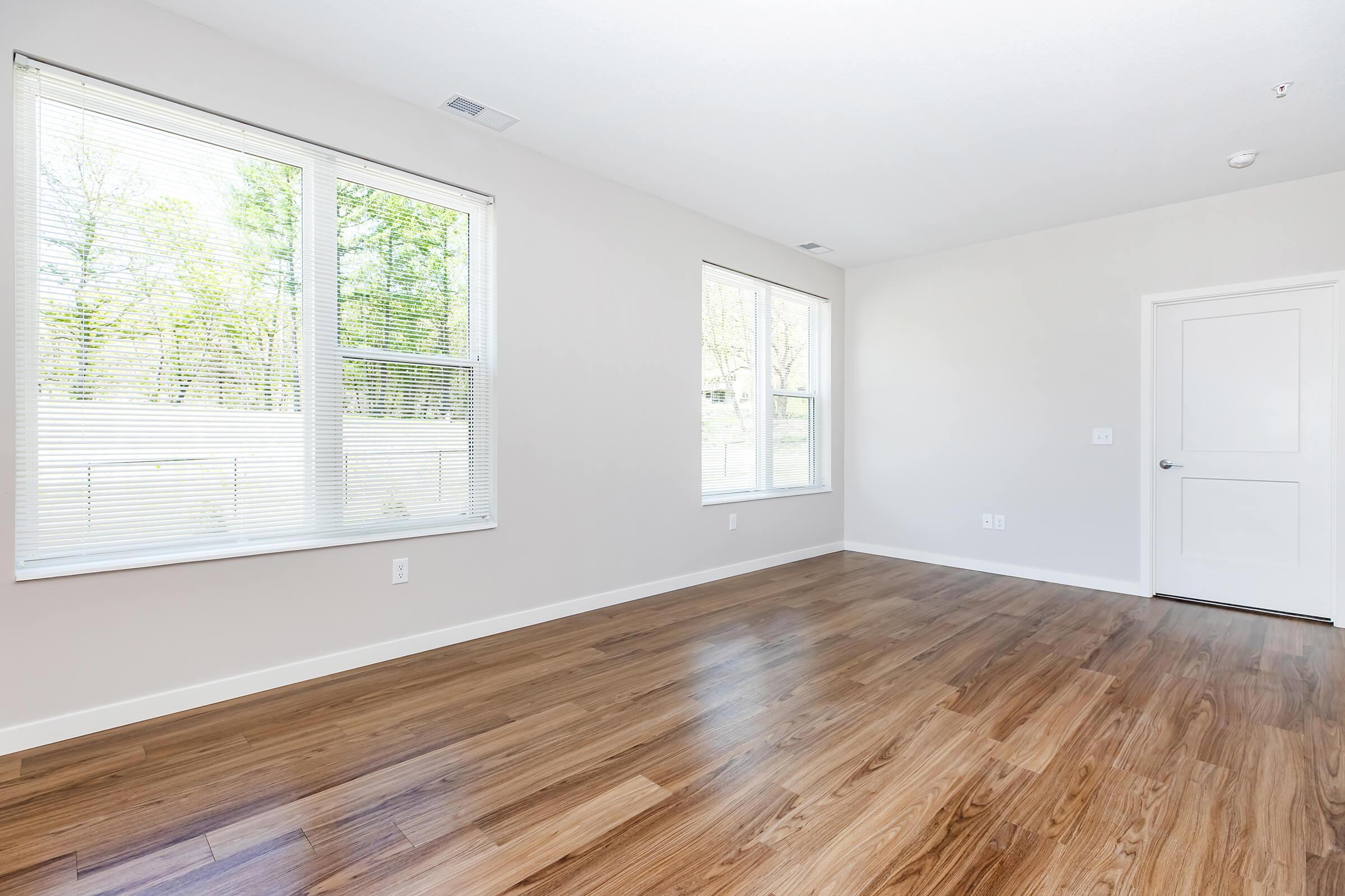 A bright, empty room with light-colored walls and hardwood flooring. There are large windows letting in natural light, and a door on the right side leads to another space. The overall atmosphere is clean and spacious, ideal for personalization or decoration.