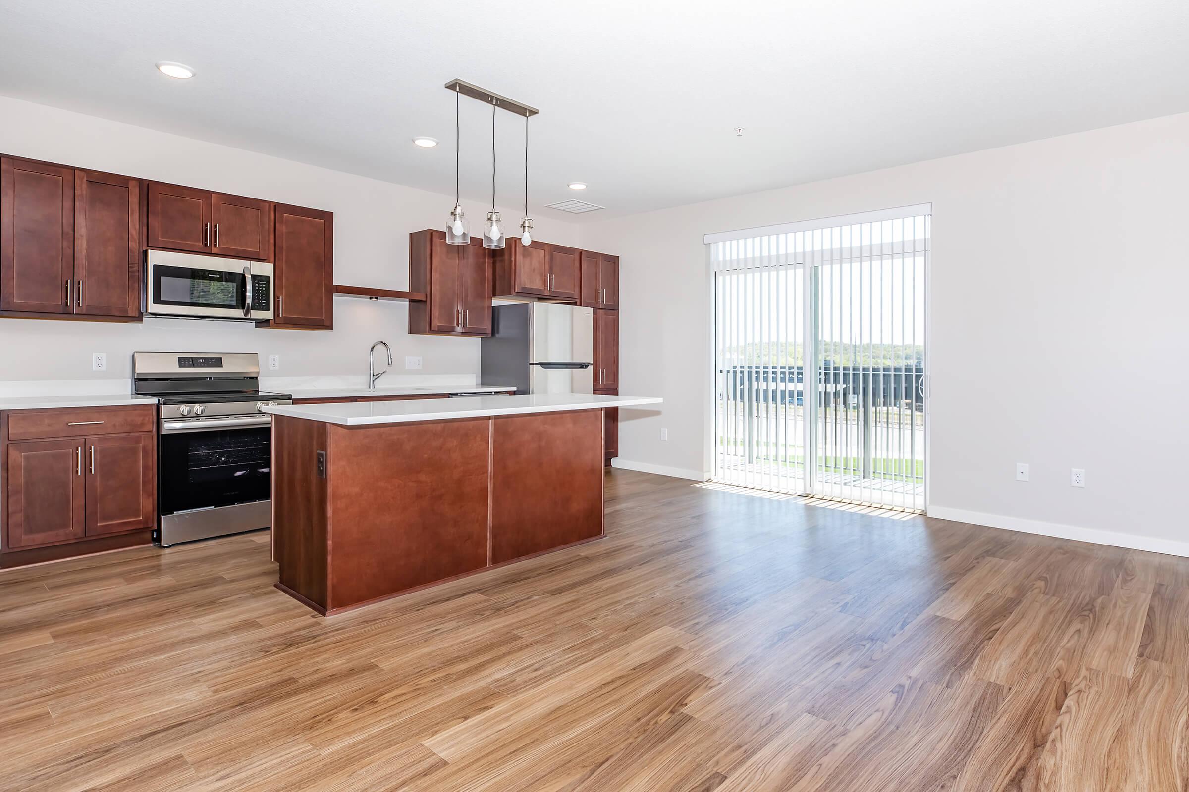 Modern kitchen with dark wood cabinetry, stainless steel appliances, and a large island. The open layout features light wooden flooring and a bright area with sliding glass doors leading to a balcony. Natural light fills the space, creating an inviting atmosphere.