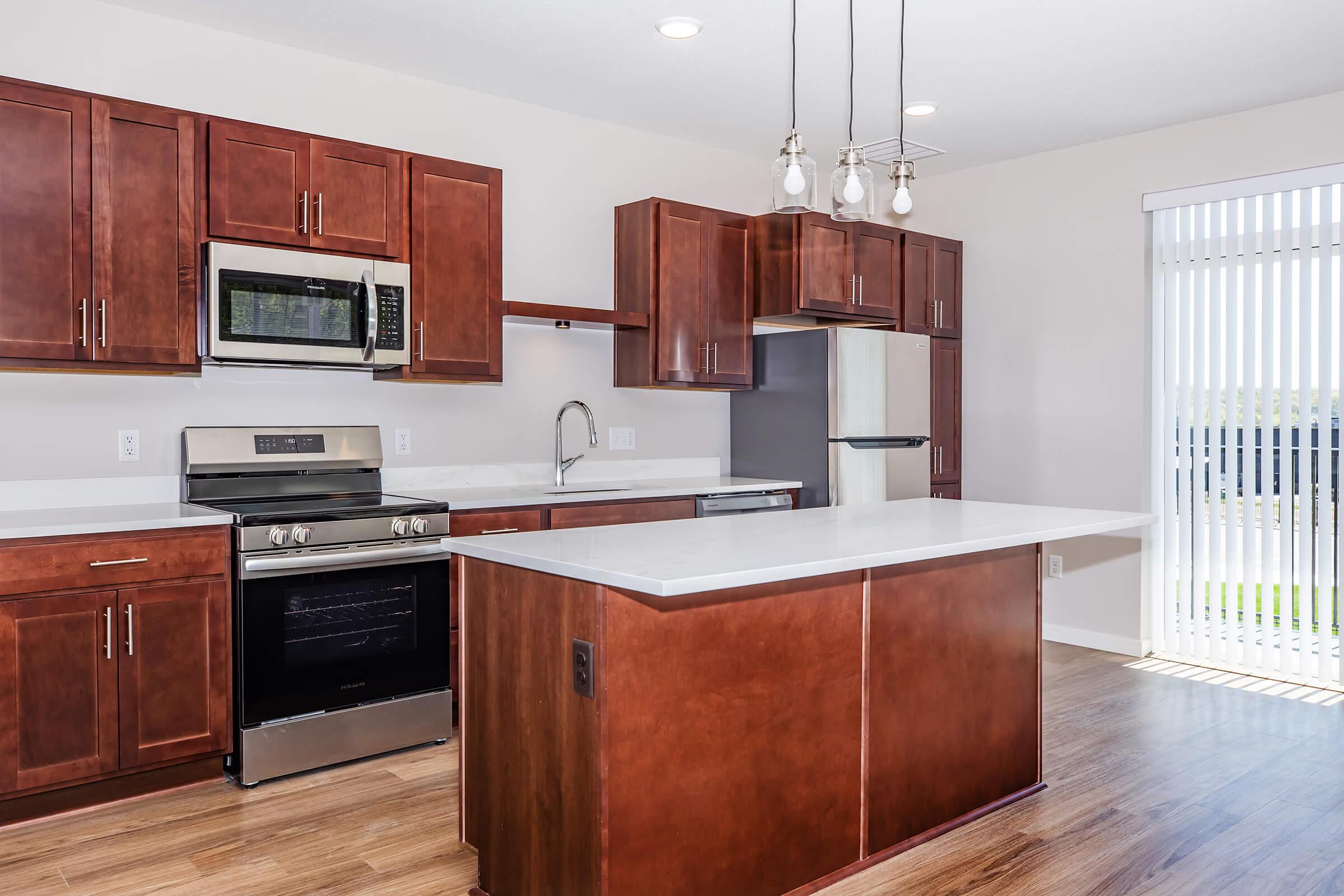 A modern kitchen featuring dark wood cabinetry, stainless steel appliances, and a white countertop. The kitchen includes a microwave, oven, and dishwasher. There's a large island in the center and vertical blinds on a window, allowing natural light to enter the space. The flooring is made of light wood.