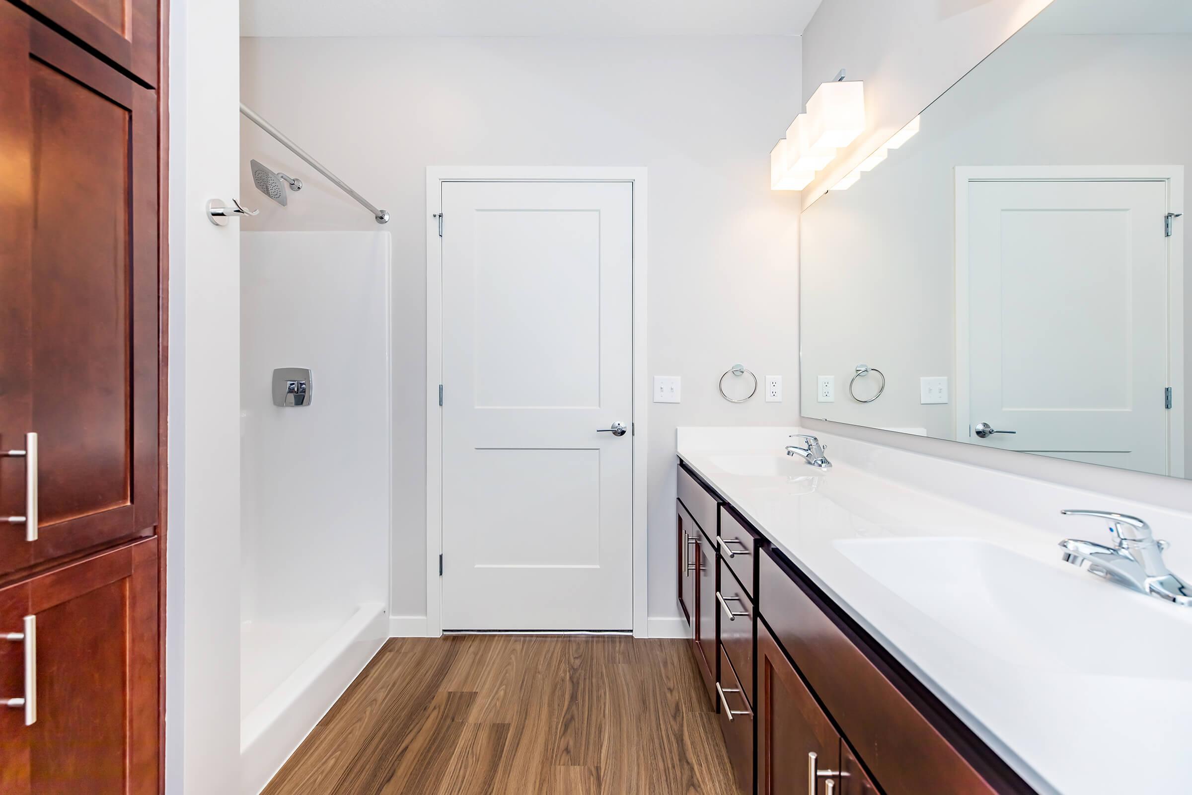 A modern bathroom featuring a single vanity with dual sinks and dark wood cabinetry, a large mirror above the sink, a walk-in shower with a glass door, and a white door leading out. The flooring is wooden, and there are contemporary light fixtures above the mirror. The walls are painted in a light color.