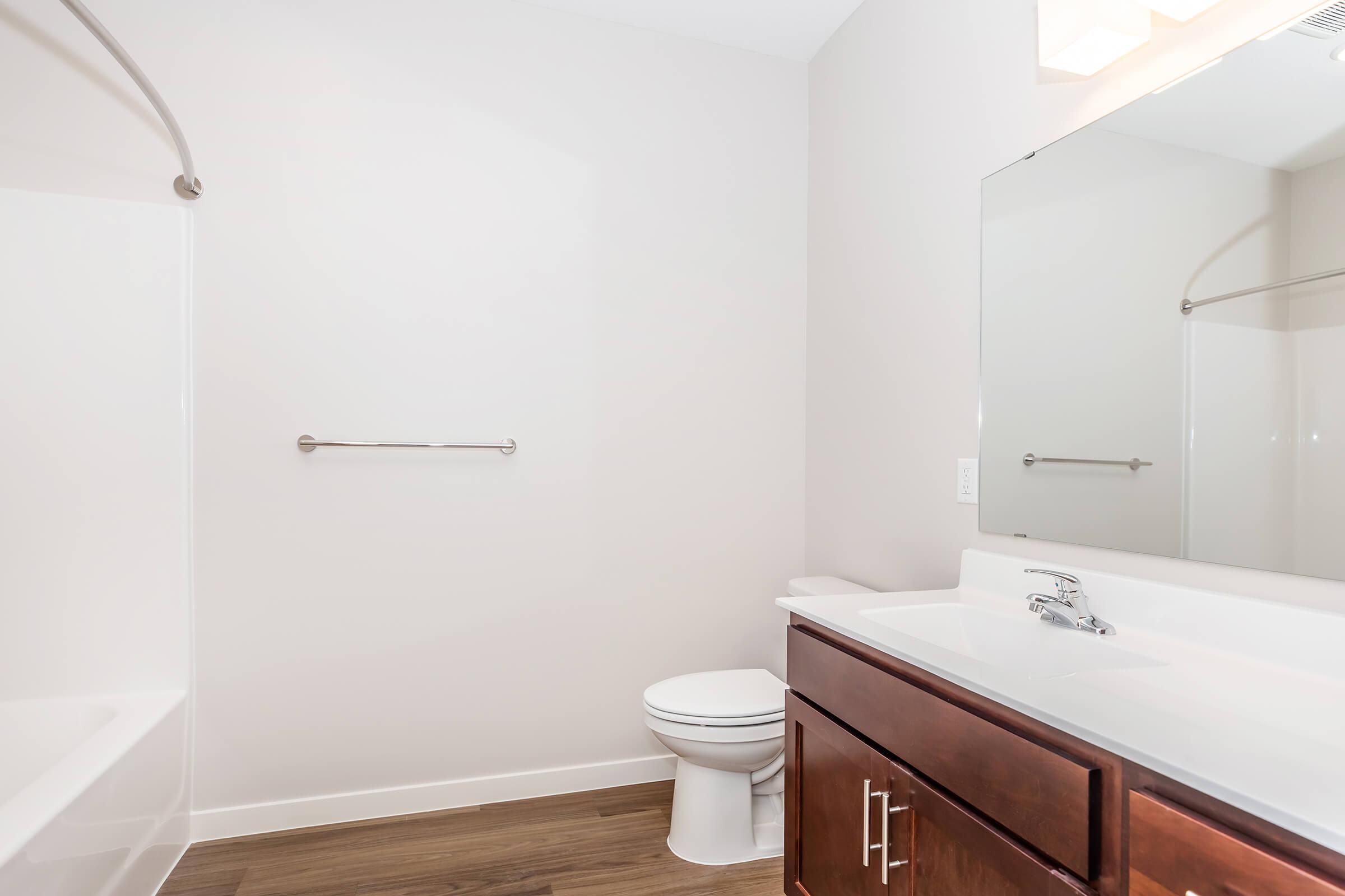 A clean, modern bathroom featuring a white bathtub, a toilet, and a wooden vanity with a sink. There is a large mirror above the sink and a towel bar on the wall. The walls are painted in a light color, and the floor has wood-like tiles, creating a bright and spacious appearance.