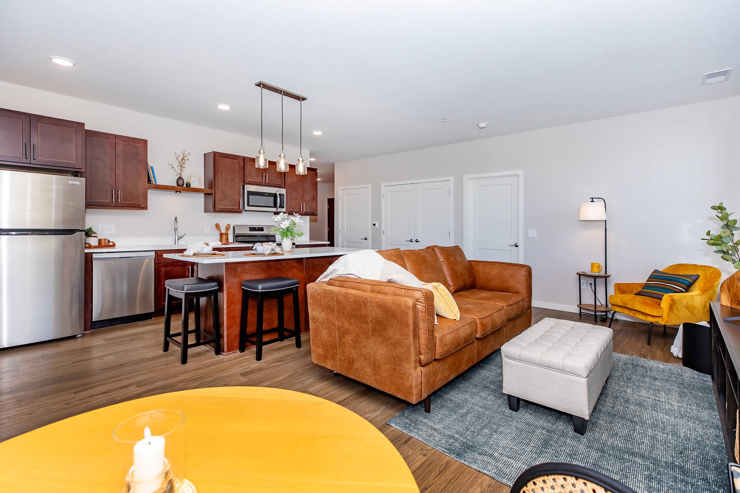 A cozy modern living space featuring a brown leather couch, a round yellow coffee table, and a gray area rug. The kitchen is equipped with stainless steel appliances and wooden cabinets. Two bar stools are positioned at the kitchen island, and additional seating is present in the adjacent area. Bright and inviting atmosphere.