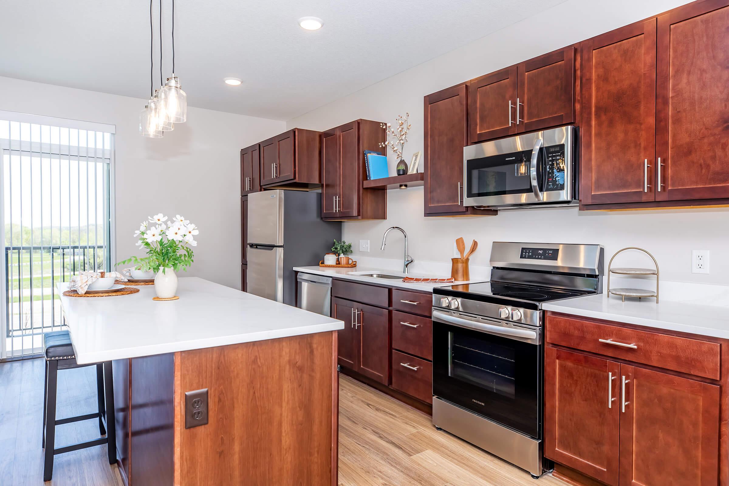 Modern kitchen featuring dark wood cabinets, stainless steel appliances, and a large island with a white countertop. The space is bright and airy, with large windows letting in natural light. Decor includes a vase of flowers on the island and a decorative piece on the shelf.