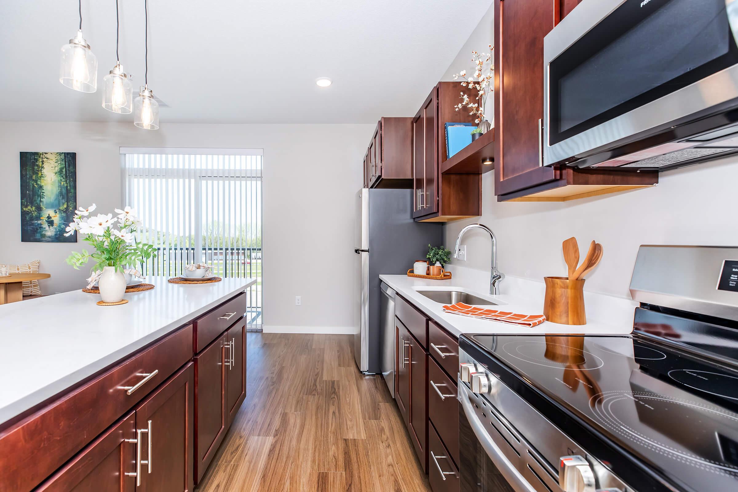 Modern kitchen featuring dark wood cabinetry, stainless steel appliances, and a large island counter. The space is well-lit with pendant lights and has a scenic view through large windows. Decorative items like a bowl of fruit and a small plant add warmth to the contemporary design.