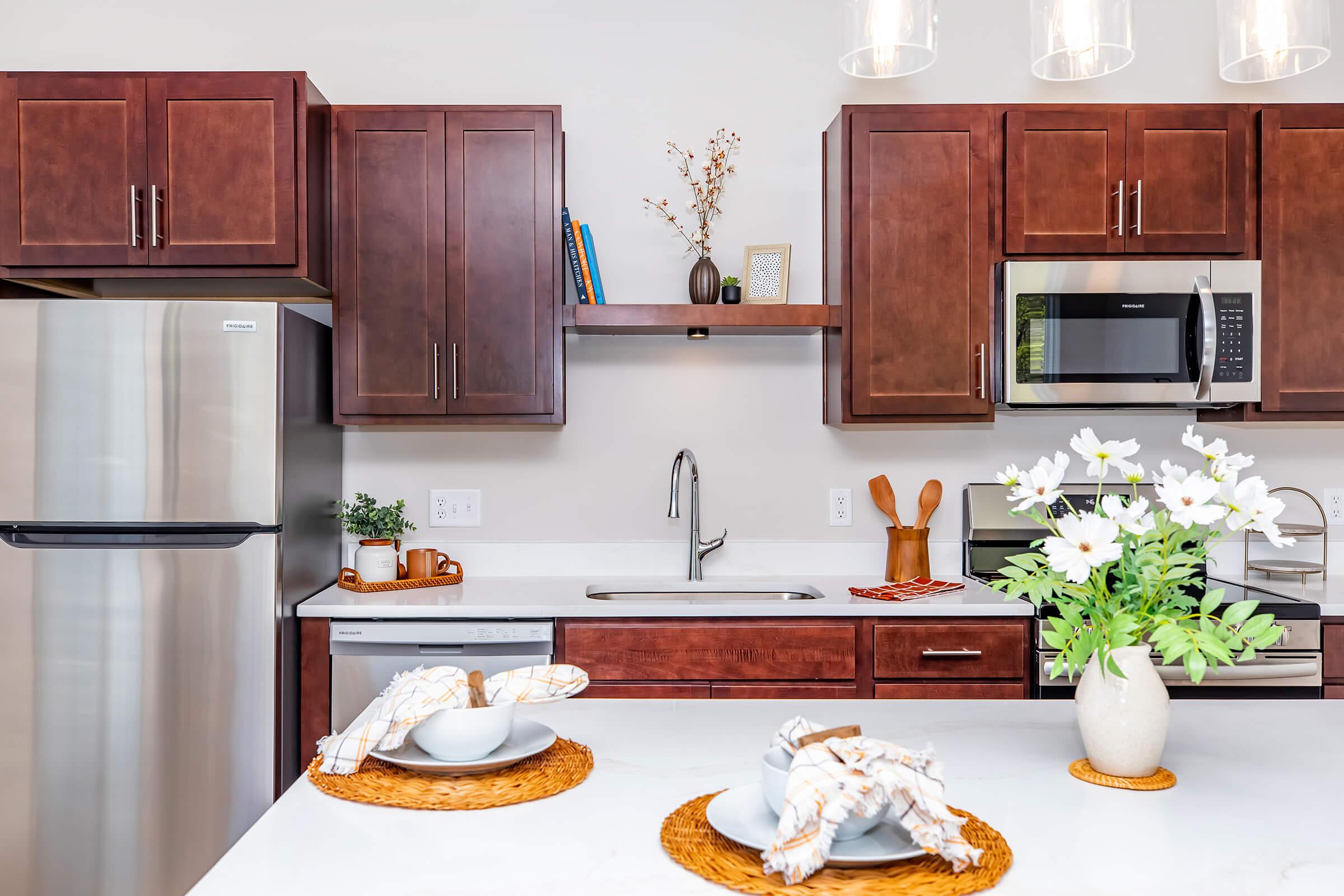 A modern kitchen featuring brown wooden cabinets, a stainless steel refrigerator, and a microwave. The countertop is adorned with a vase of white flowers and two place settings on woven placemats. A shelf above the sink holds decorative items, creating a warm and inviting atmosphere.