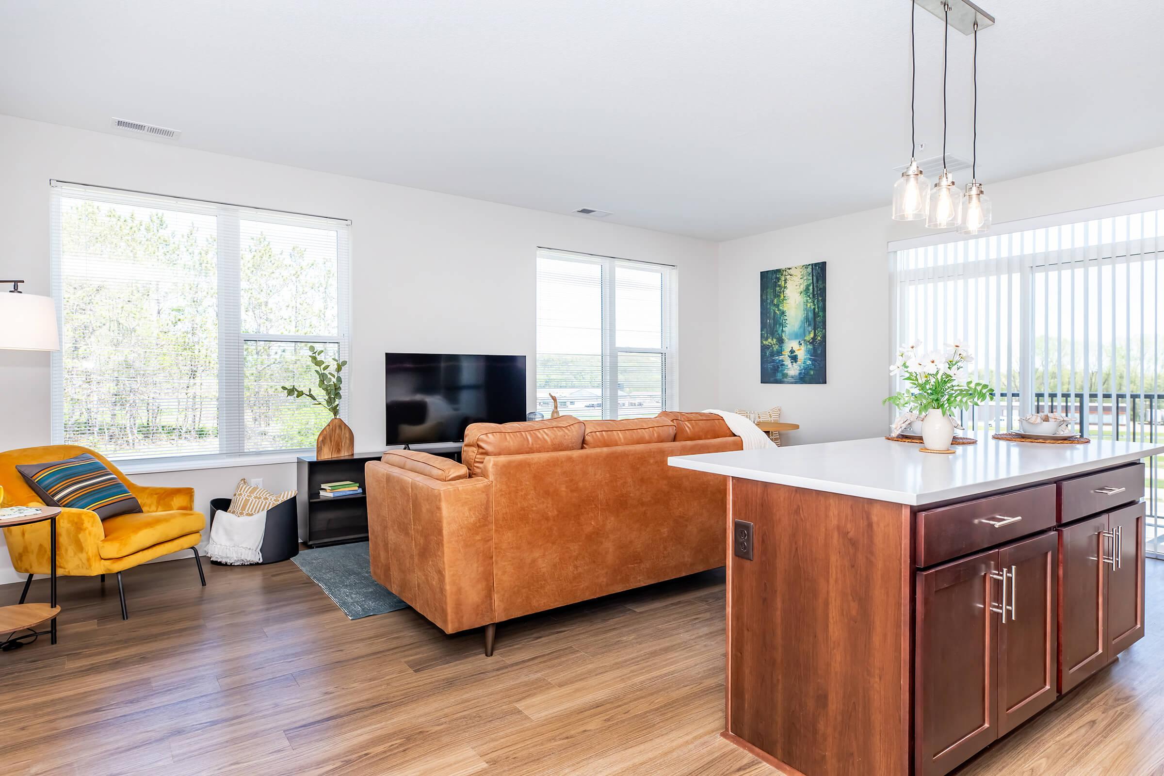 A modern living room with a brown leather sofa and a television unit. A cozy yellow chair is positioned nearby. The space has natural light from large windows and a view of greenery outside. There's a kitchen island with dark wooden cabinets and decorative plants on the countertop, creating a welcoming atmosphere.