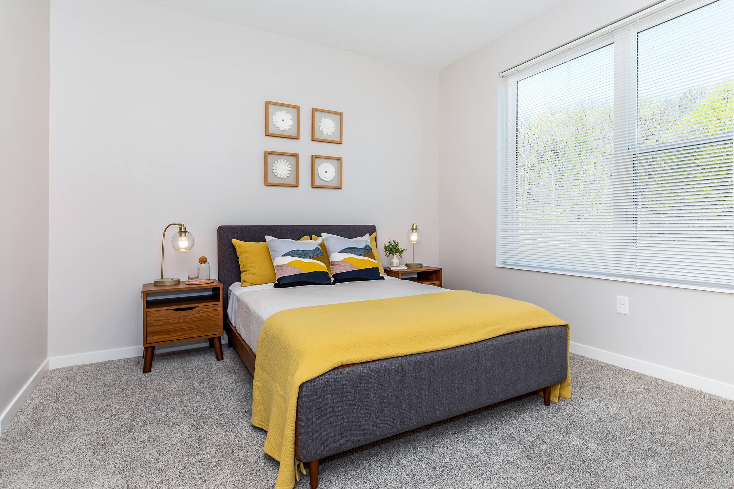 A cozy bedroom featuring a gray upholstered bed with a yellow blanket and decorative pillows. Two bedside tables with lamps are on either side of the bed. Above the bed are four framed wall art pieces. The room has large windows allowing natural light to enter, with light-colored walls and carpeted flooring.