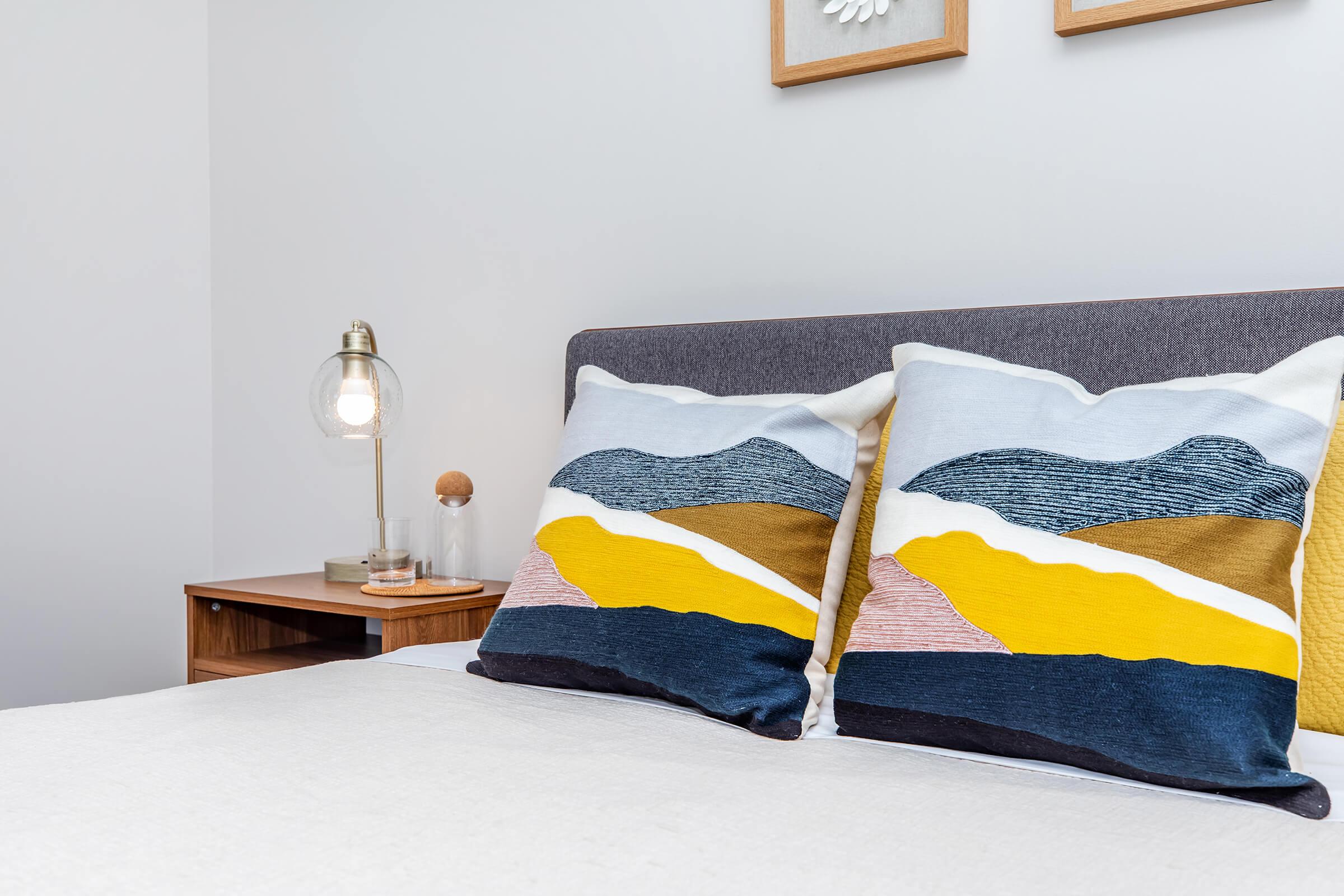 A neatly made bed with two decorative pillows featuring abstract patterns in blue, yellow, and brown. A bedside table holds a lamp with a glass bulb and a small decorative item, while framed art hangs on the wall above the bed, creating a cozy and modern bedroom atmosphere.
