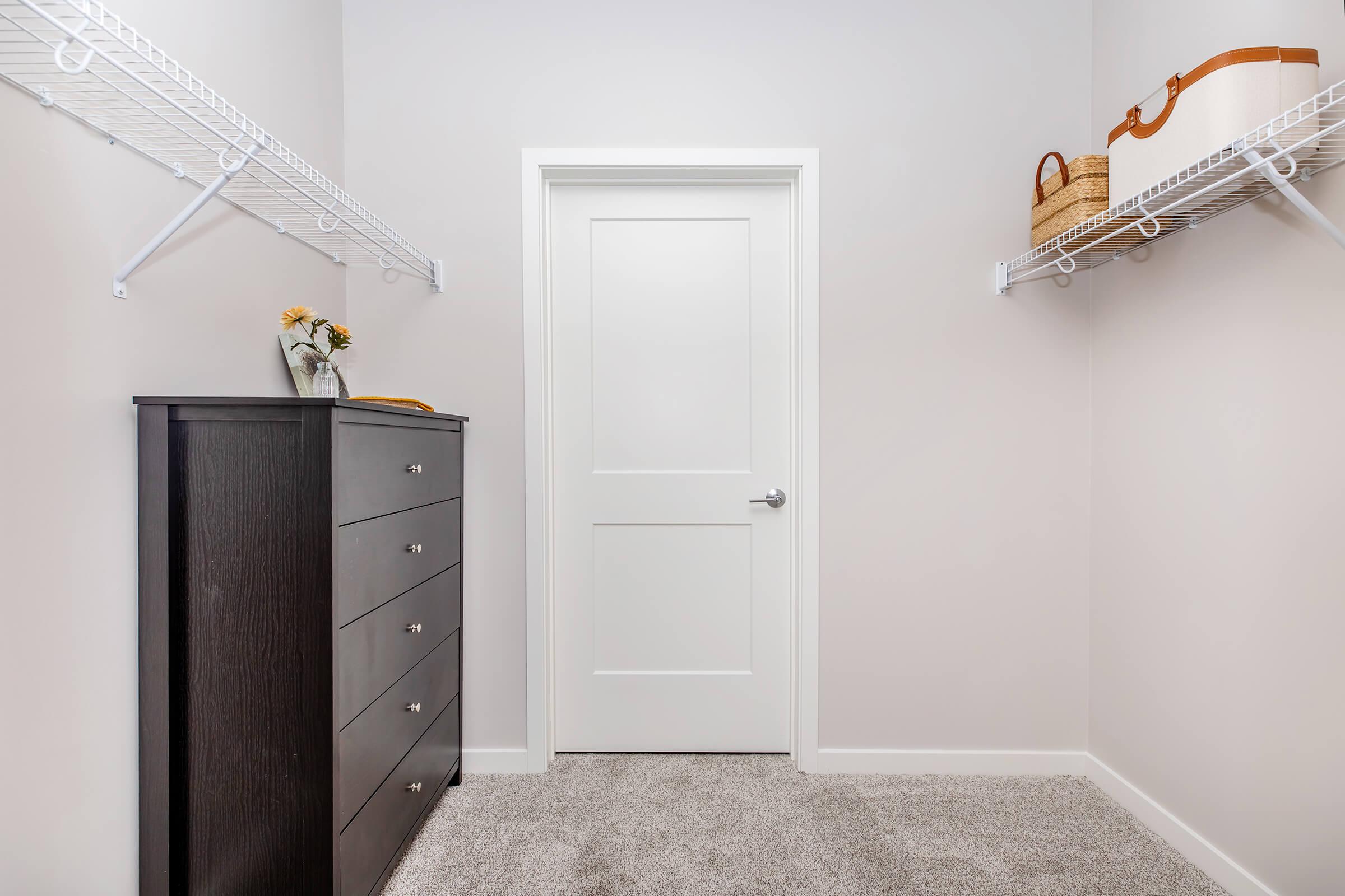 A well-organized closet featuring a dark wooden dresser with multiple drawers, two wire shelves on either side, and a closed white door. Decorative items, including a vase with flowers and a wicker basket, adorn the shelves. The floor is carpeted, and the walls are painted a light color.