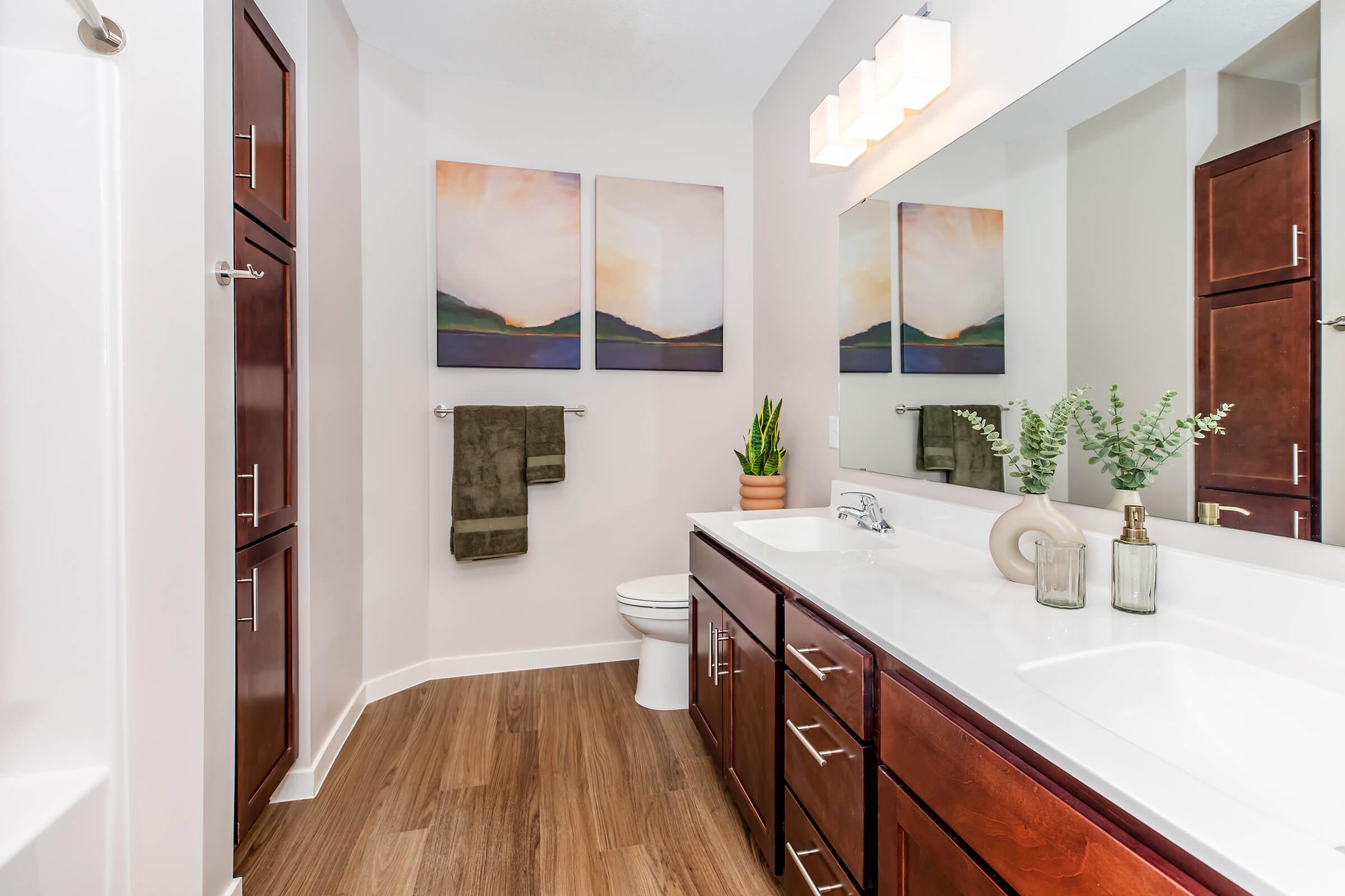 Modern bathroom featuring a double vanity with dark wood cabinetry, a large mirror, and decorative plants. Two abstract paintings hang above the sink and neatly arranged towels are visible on a towel rack. The space is well-lit, showcasing a combination of contemporary design and natural elements.