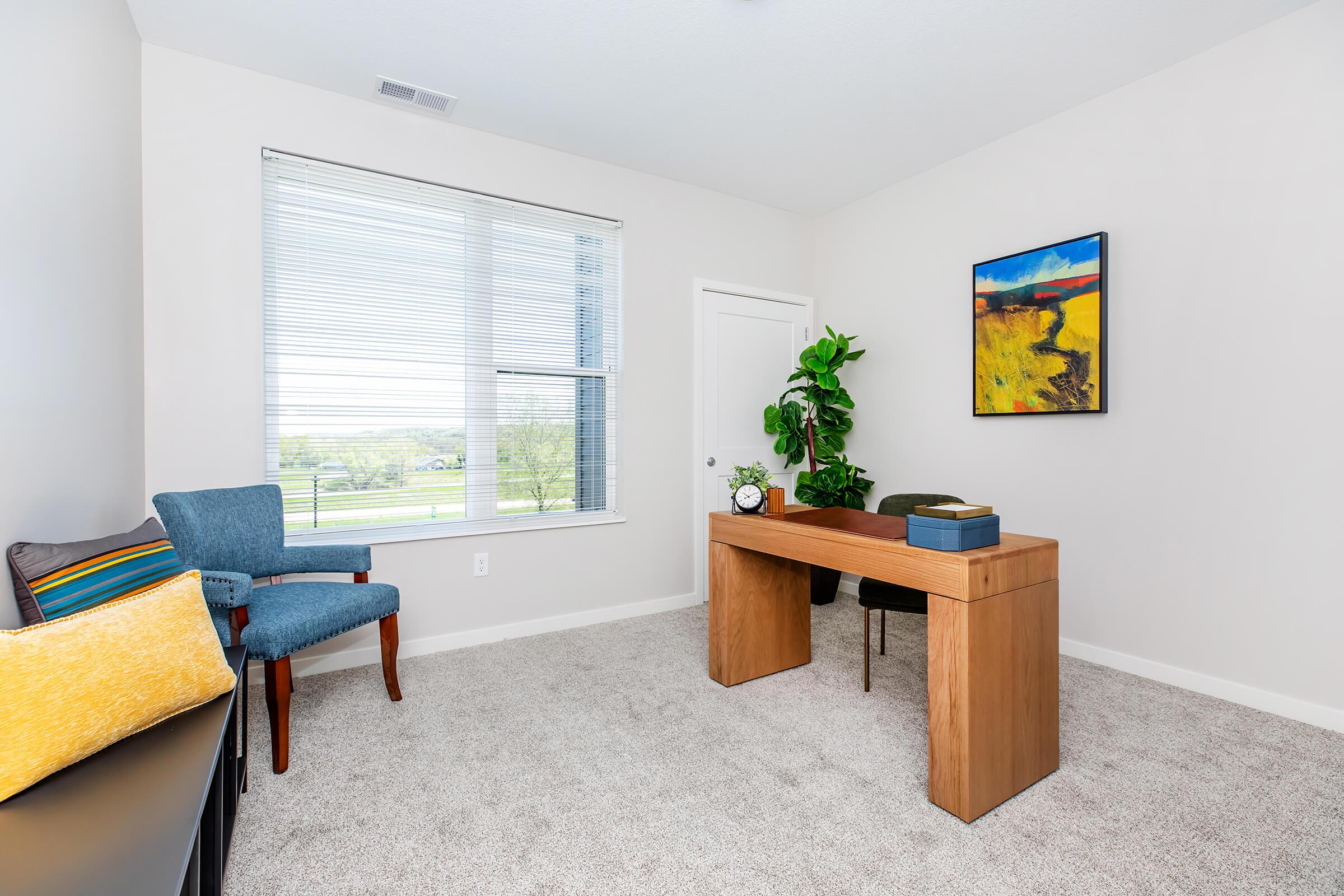 A well-lit home office featuring a wooden desk, a blue chair, and a decorative plant. There’s a colorful painting on the wall and a cozy couch with colorful cushions. Natural light streams in through a window with blinds, creating a bright and inviting workspace. The room has neutral walls and a plush carpet.