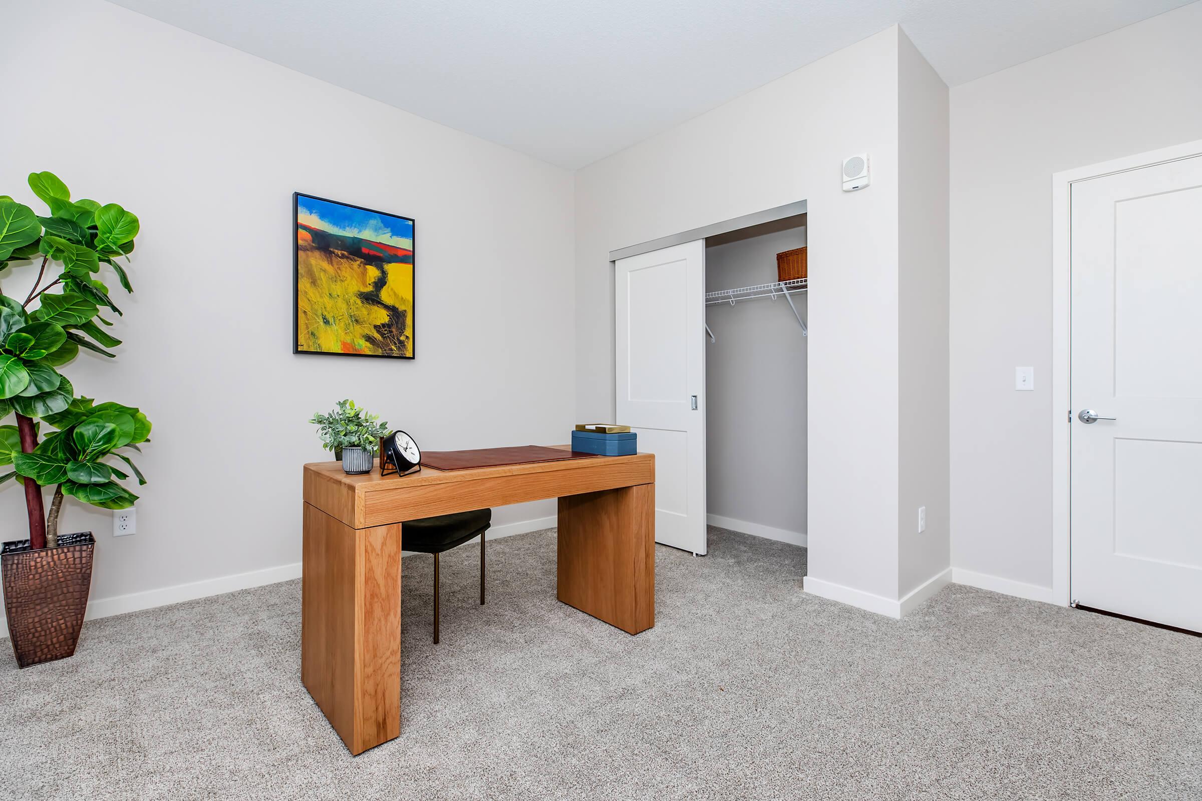A modern home office featuring a wooden desk with a clock and a small decorative plant. The room is carpeted and has neutral-colored walls. There's a framed artwork on the wall, a closet with sliding doors, and a single black chair positioned at the desk. A tall potted plant is in the corner.