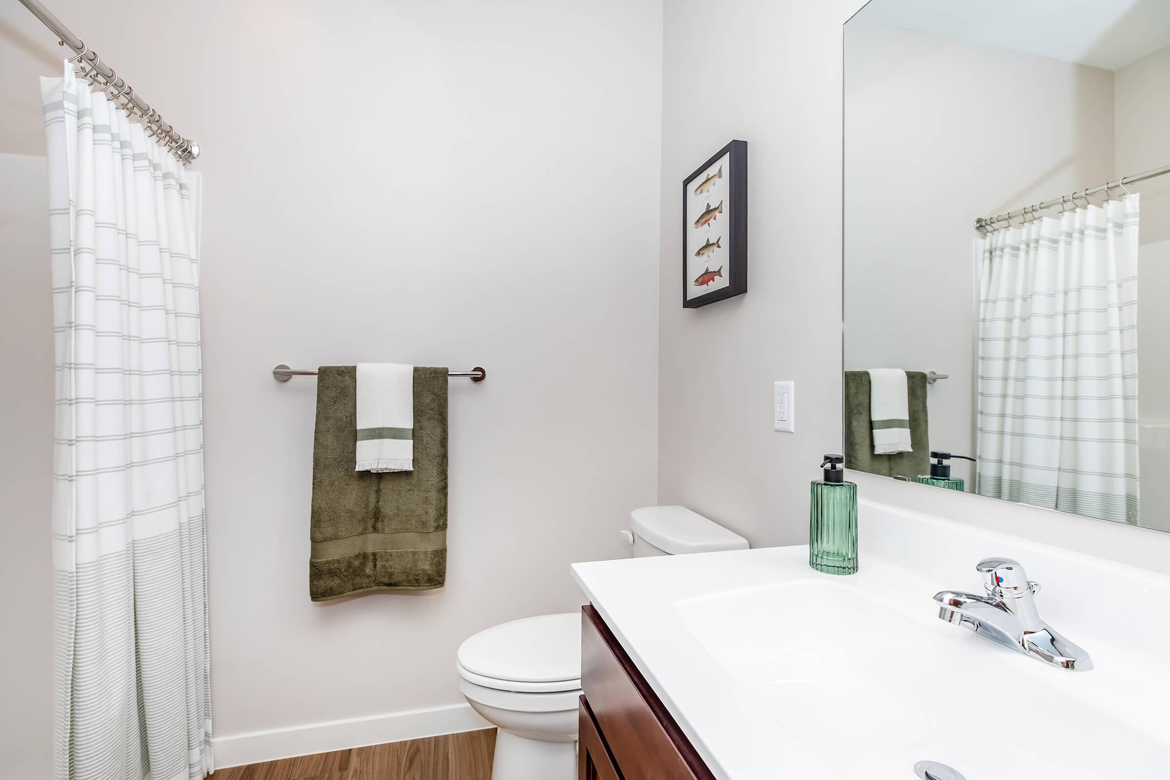 A modern bathroom featuring a shower with a white and gray striped curtain, a white toilet, and a clean vanity with a sink. Above the sink, there's a rectangular mirror and a wall decoration showcasing images of fish. A green hand towel hangs on a rack, adding a touch of color to the neutral palette.