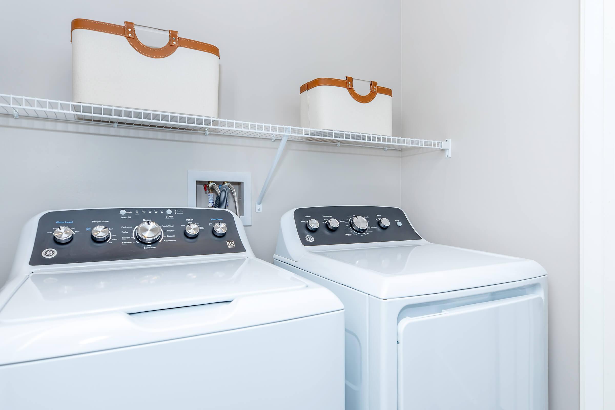 A laundry room featuring a white washing machine and dryer with control panels on top. Above them, a wire shelf holds two storage baskets with tan leather handles. The walls are painted light gray, creating a clean and tidy appearance.