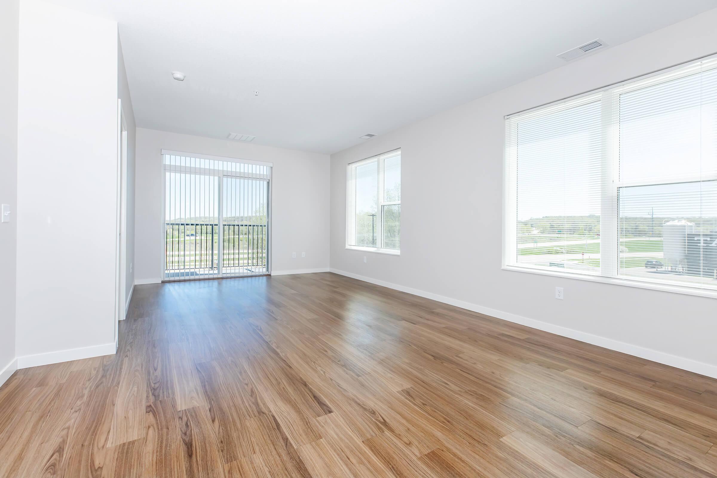 A spacious, empty living room featuring light-colored walls and large windows allowing natural light. The floor is made of wooden laminate, and there are sliding glass doors leading to a balcony. The overall atmosphere is bright and open, ideal for furnishing and decorating.