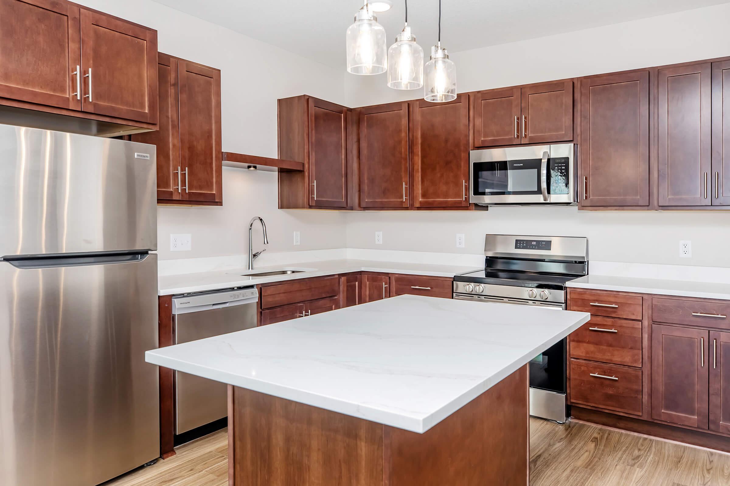 Modern kitchen featuring dark wood cabinetry, stainless steel appliances, and a large island with a white countertop. The kitchen includes a refrigerator, oven, microwave, and dishwasher, with pendant lighting above the island and a neutral wall color.