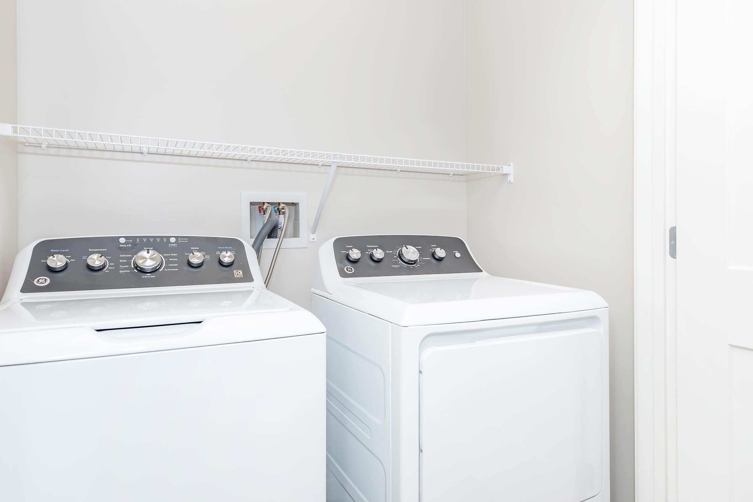 A laundry room featuring a washing machine and a dryer side by side. Both appliances have modern designs with digital controls. A white wire shelf is mounted above them for storage, and the walls are a light color, creating a clean and organized space. A doorway is visible on the right.