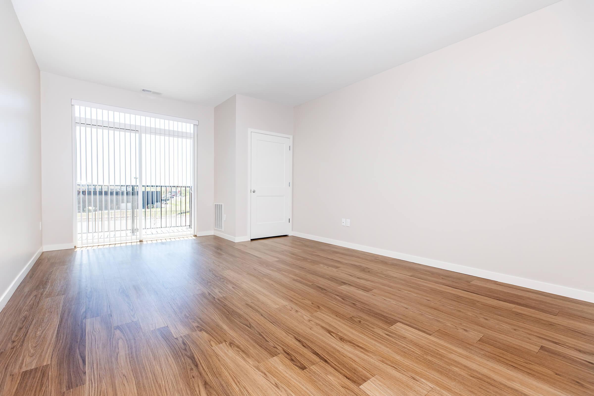 Spacious empty room with light-colored walls and wooden flooring, featuring a sliding glass door with vertical blinds leading to a balcony, and a closed door on the side. Natural light illuminates the space, creating a bright and airy atmosphere.