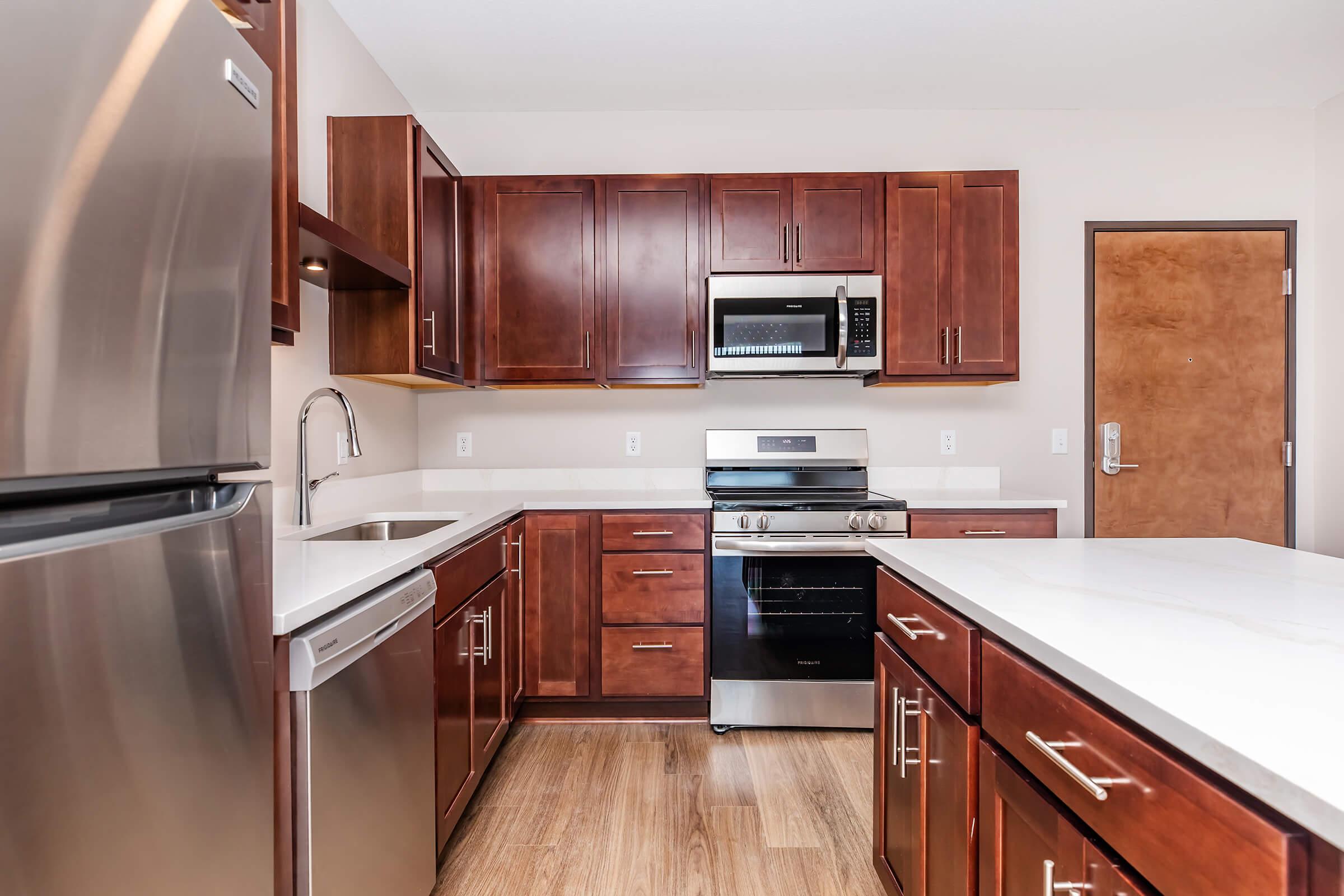 Modern kitchen featuring dark wood cabinetry, stainless steel appliances including a refrigerator, microwave, and oven. The countertop is white with a sink installed. The flooring is light wood, creating a warm and inviting atmosphere. The layout is open and functional, ideal for cooking and entertaining.