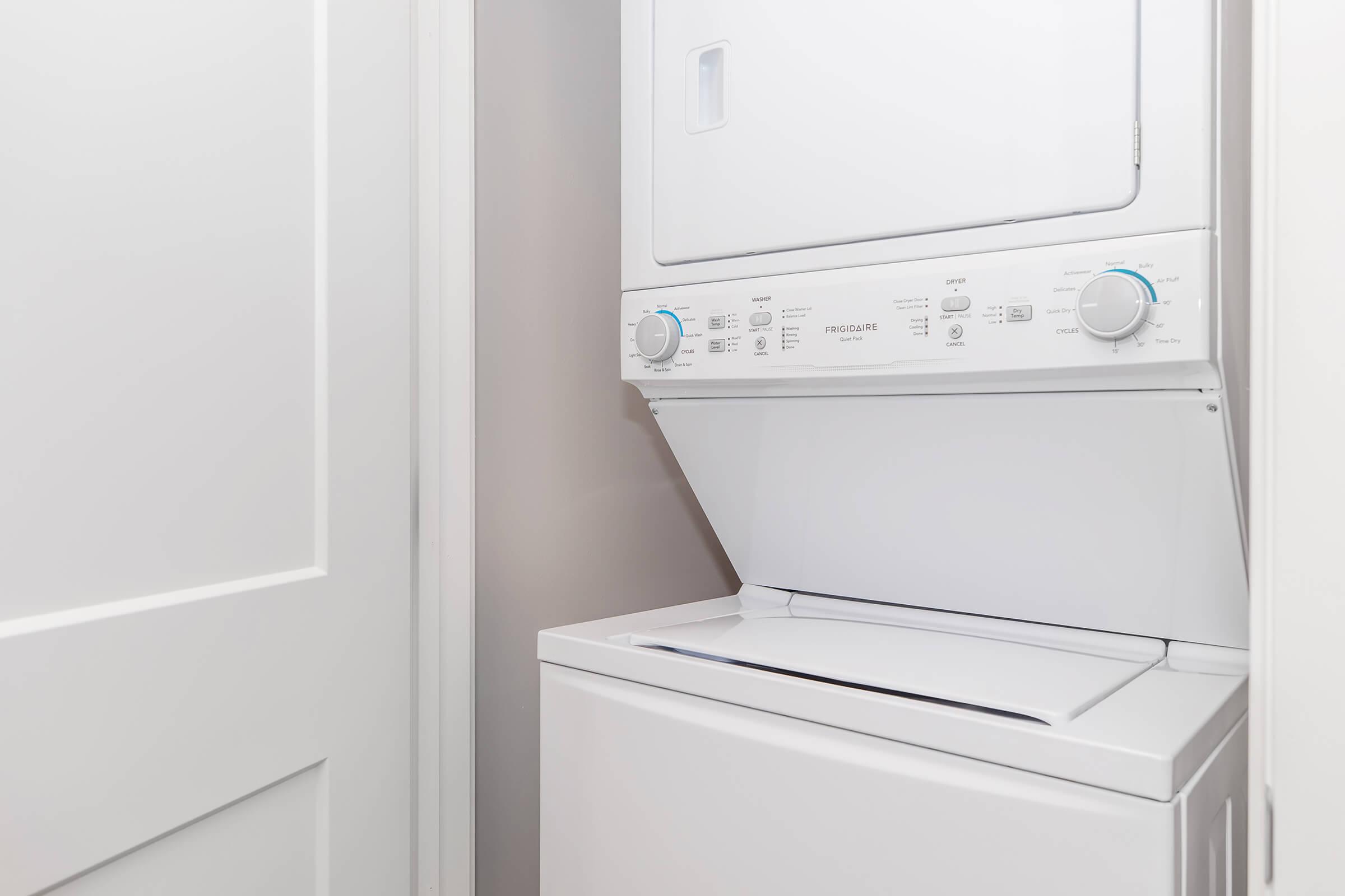 A stacked washer and dryer set in a laundry room corner, featuring a white exterior with control knobs and buttons. The appliance is positioned next to a closed white door, against a light-colored wall.