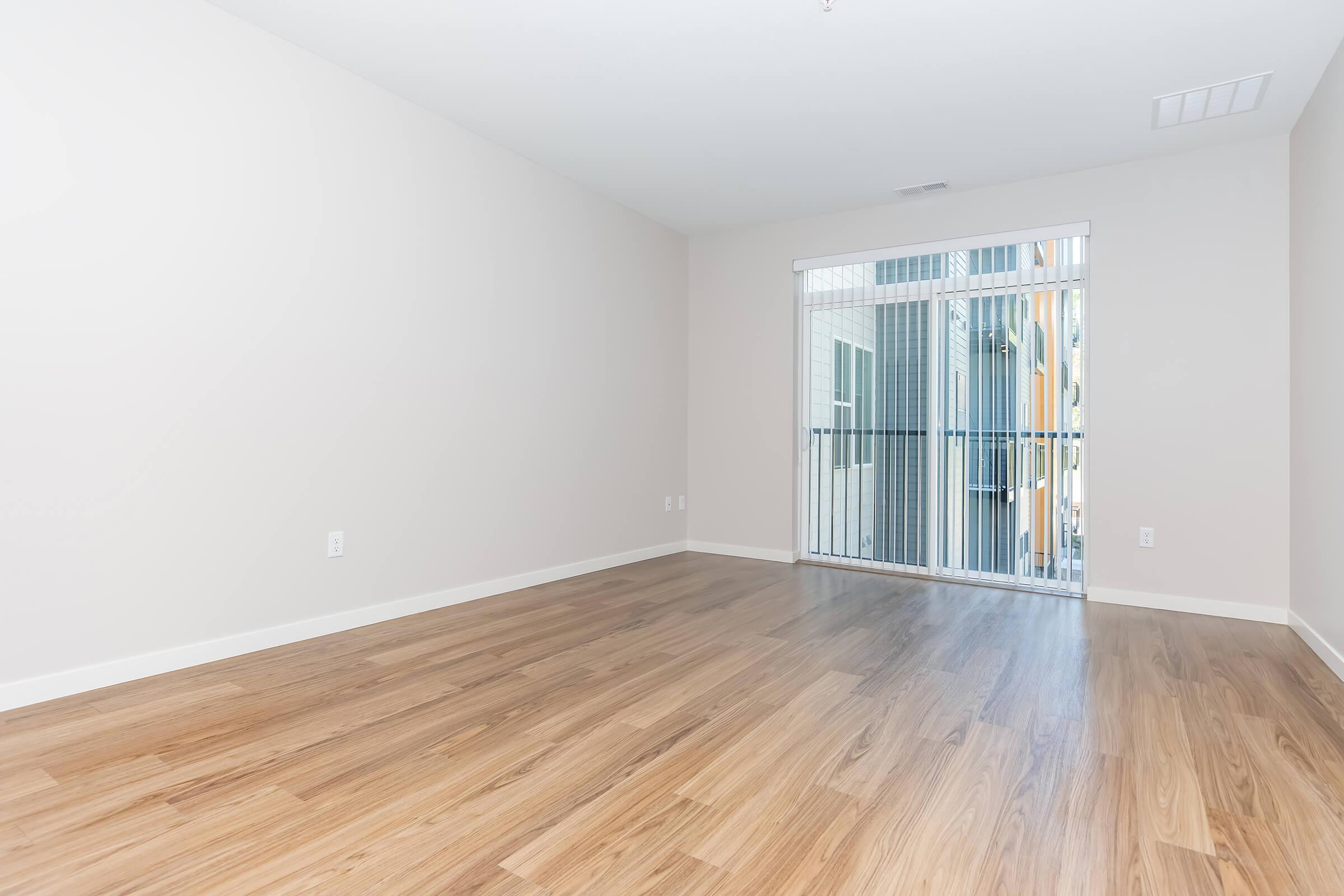 Spacious, empty room with light wood flooring and neutral-colored walls. A large sliding glass door leads to a balcony with a view of a multi-colored building exterior. The room is bright and airy, with natural light coming in through the door.