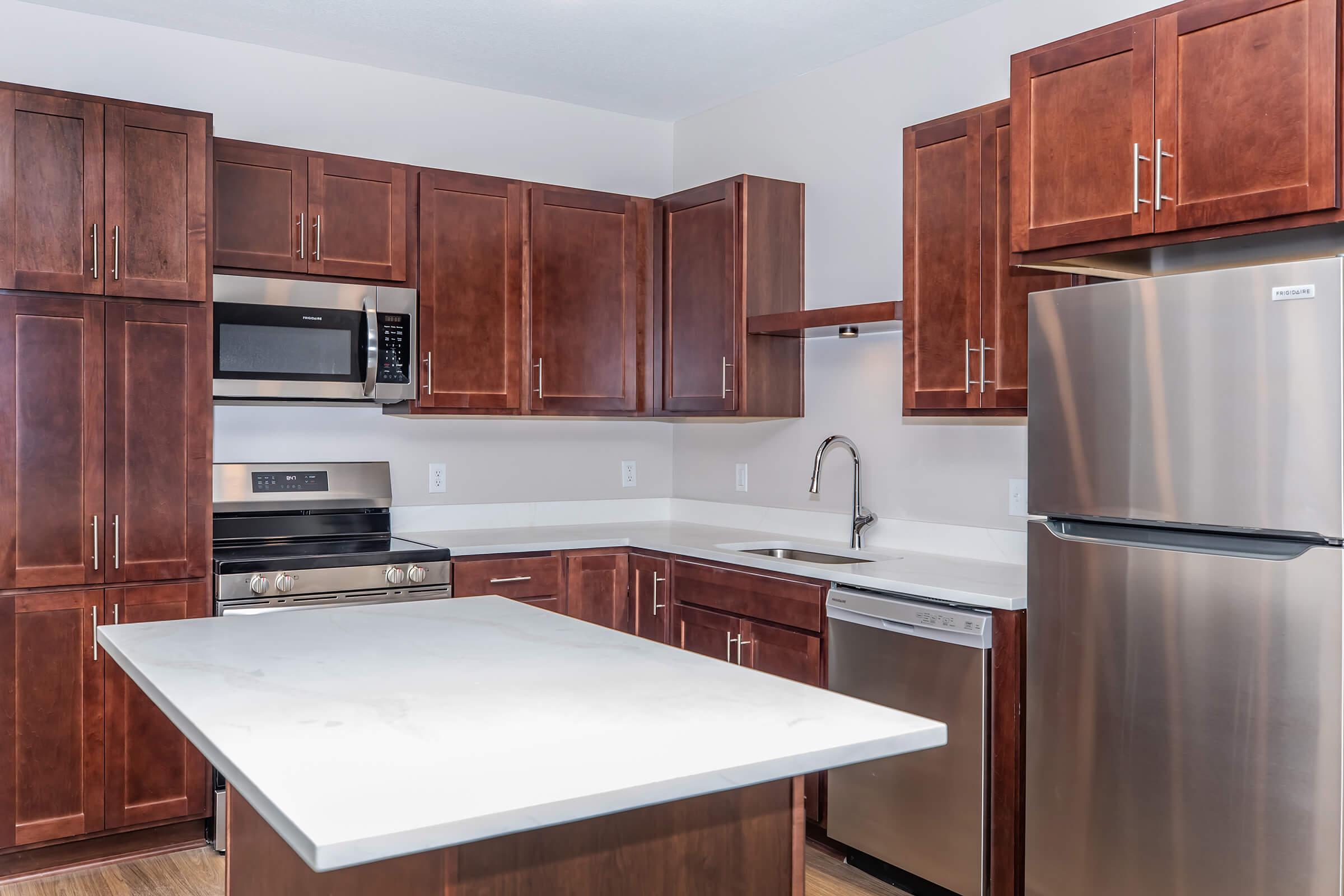Modern kitchen featuring dark wood cabinetry, stainless steel appliances including a refrigerator and oven, a microwave, and a dishwasher. The white countertop contrasts with the wood, and there's an island in the center for additional workspace. Bright lighting enhances the open feel of the space.