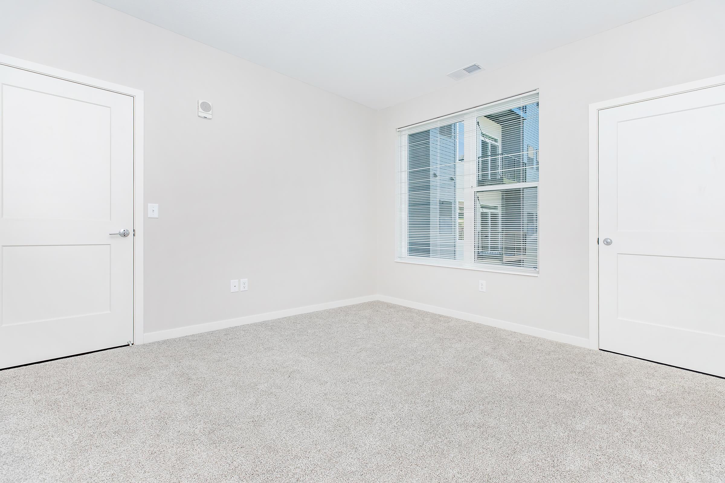A bright, empty room with light beige carpet, featuring two closed doors on the left and a large window on the right. The window has white blinds and overlooks an outdoor area with a balcony visible in the background. The walls are painted in a light, neutral color, creating a spacious atmosphere.