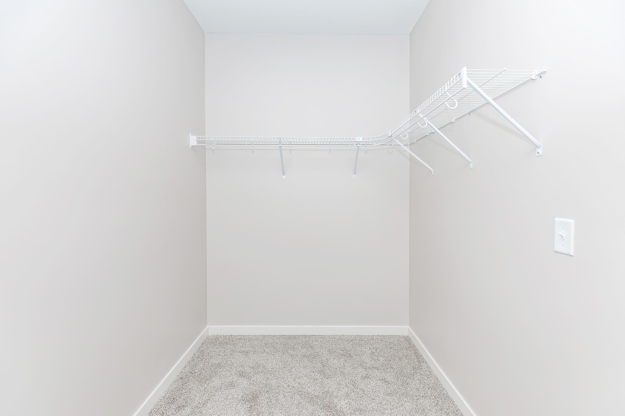 Empty room with light gray walls and a carpeted floor. There is a white wire shelf mounted on one wall, extending to one side, with no items stored on it. The space is devoid of furniture or decoration, creating a minimalistic and open feel.