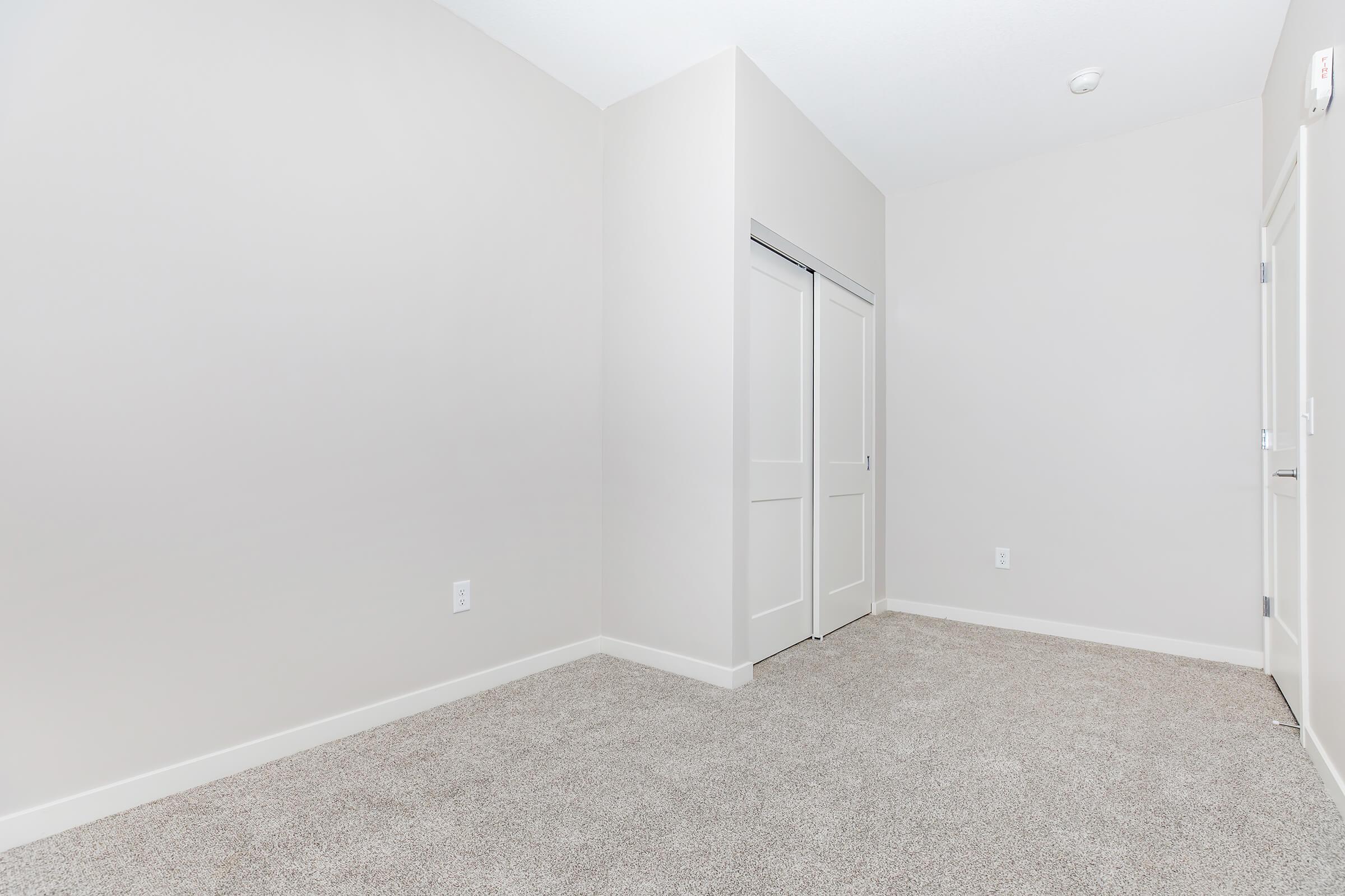 Empty room with light gray walls and beige carpet. There are two closet doors on one side and a white door leading to another area. The space is well-lit and devoid of furniture or decorations, giving a clean and minimalistic appearance.