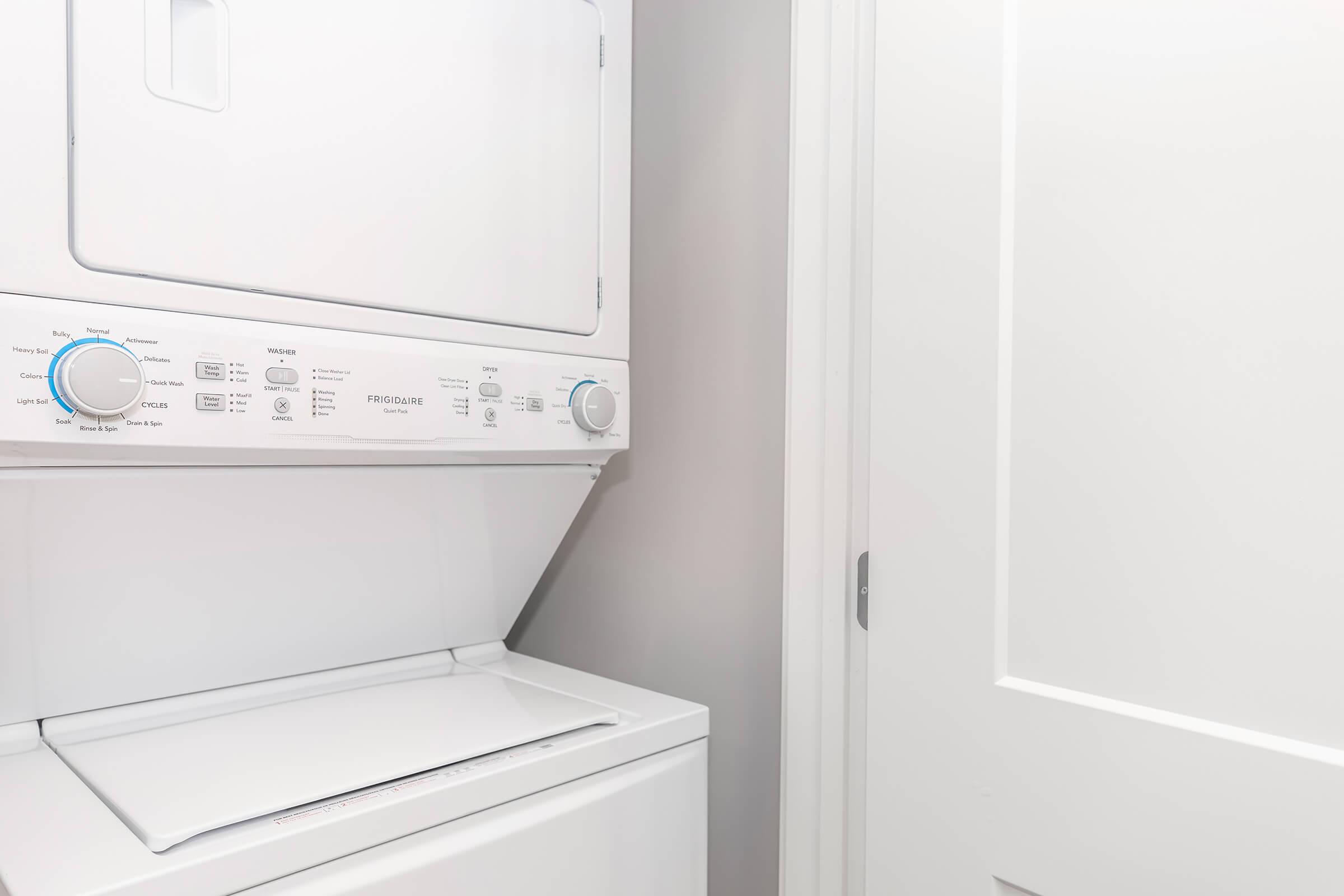 A stacked washer and dryer unit in a small laundry space. The appliance is white and features multiple control knobs and buttons. A closed white door is visible to the right, suggesting a freshly painted or modern interior setting.