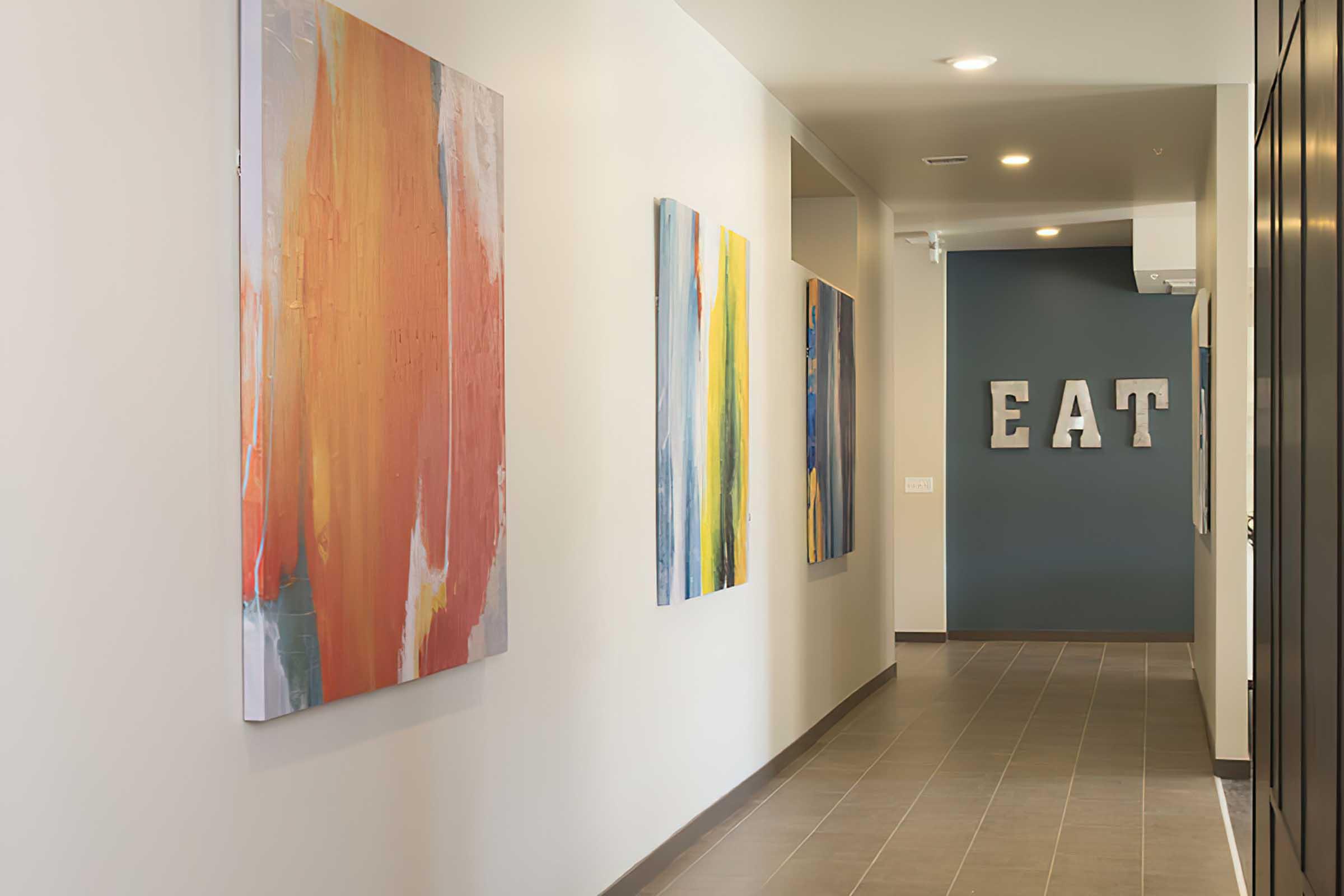 A modern hallway featuring abstract paintings in warm and cool colors, with a dark accent wall that has the letters "EAT" displayed prominently.