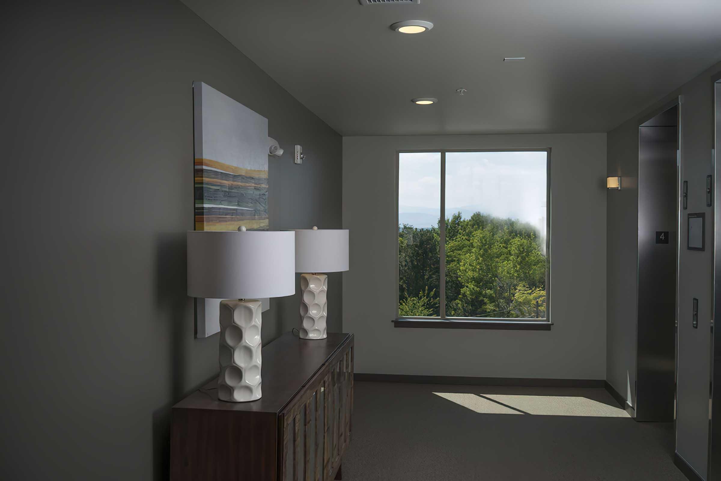 A modern hallway featuring a console table with two white lamps, a large piece of abstract artwork on the wall, and a large window that lets in natural light, showcasing green trees outside. An elevator door is visible on the right side.