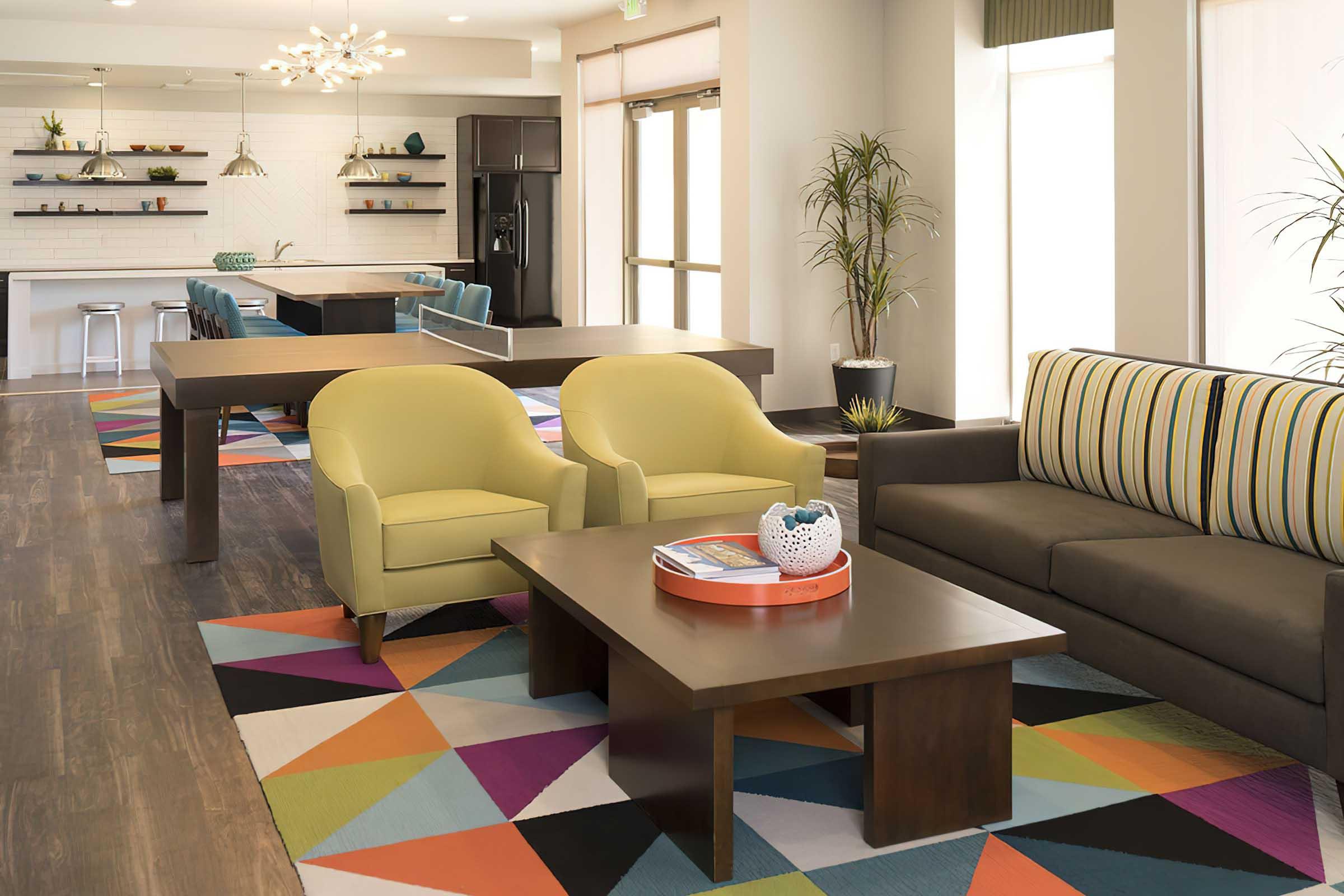 A modern lounge area featuring two bright yellow armchairs, a dark sofa with striped cushions, and a wooden coffee table. The floor has a colorful geometric rug, and there are several green plants.