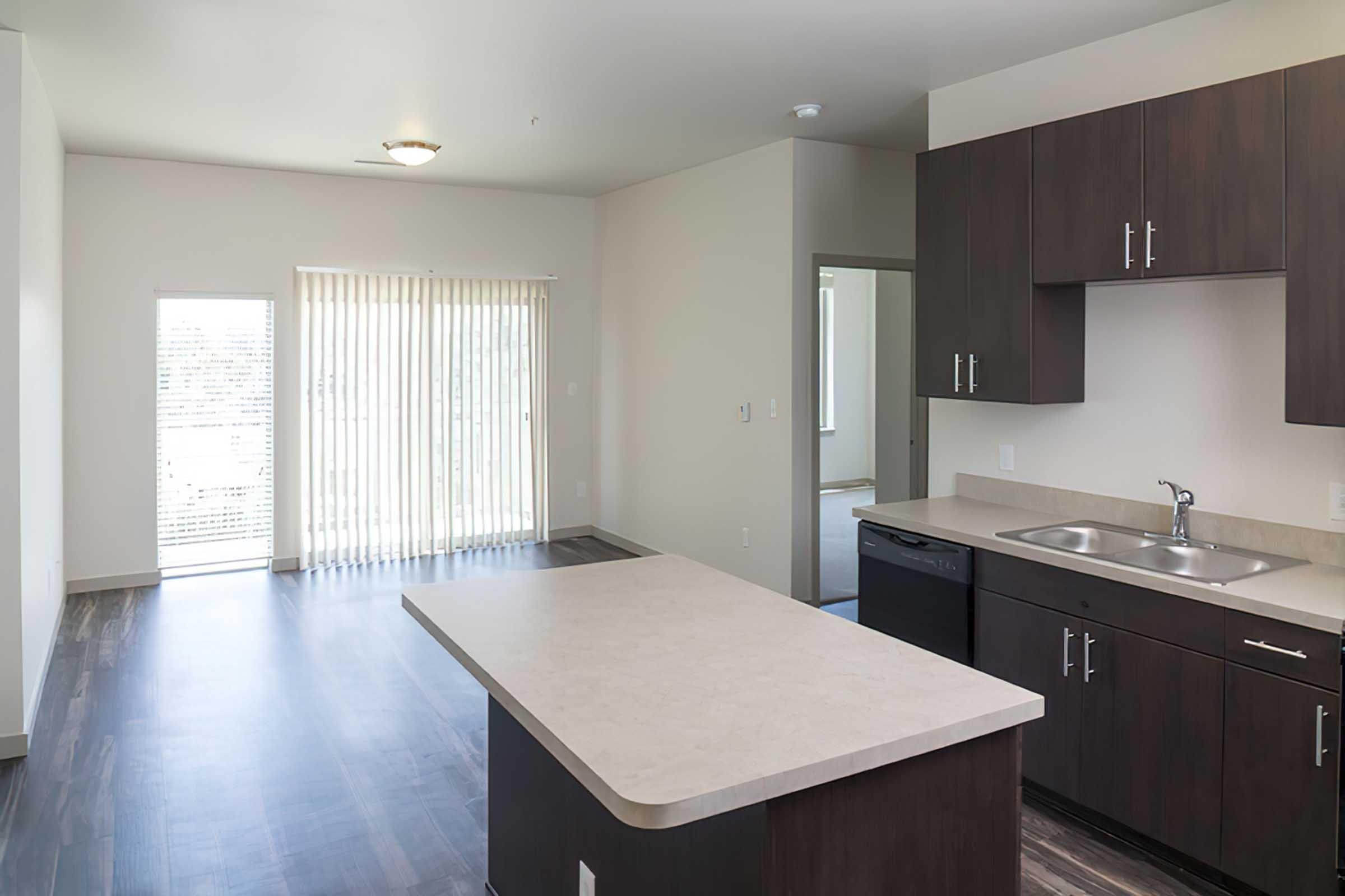 A modern kitchen with dark cabinetry and a countertop island opens into a spacious living area with large windows and blinds