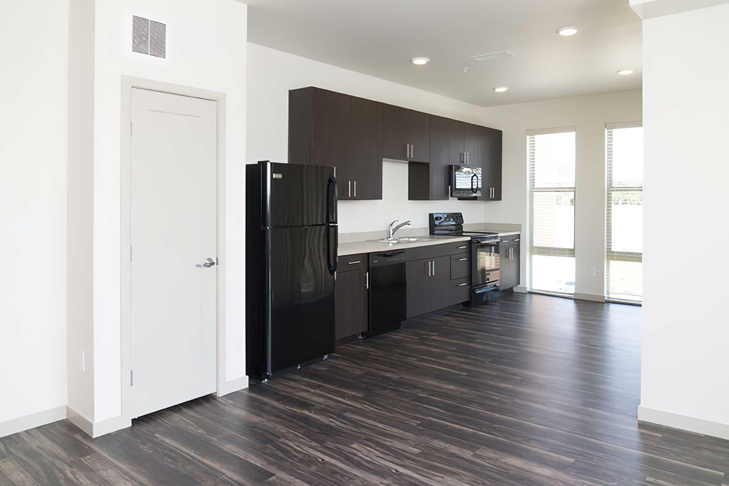 Modern kitchen in a spacious apartment with dark wood cabinetry, a black refrigerator, and large windows allowing natural light.