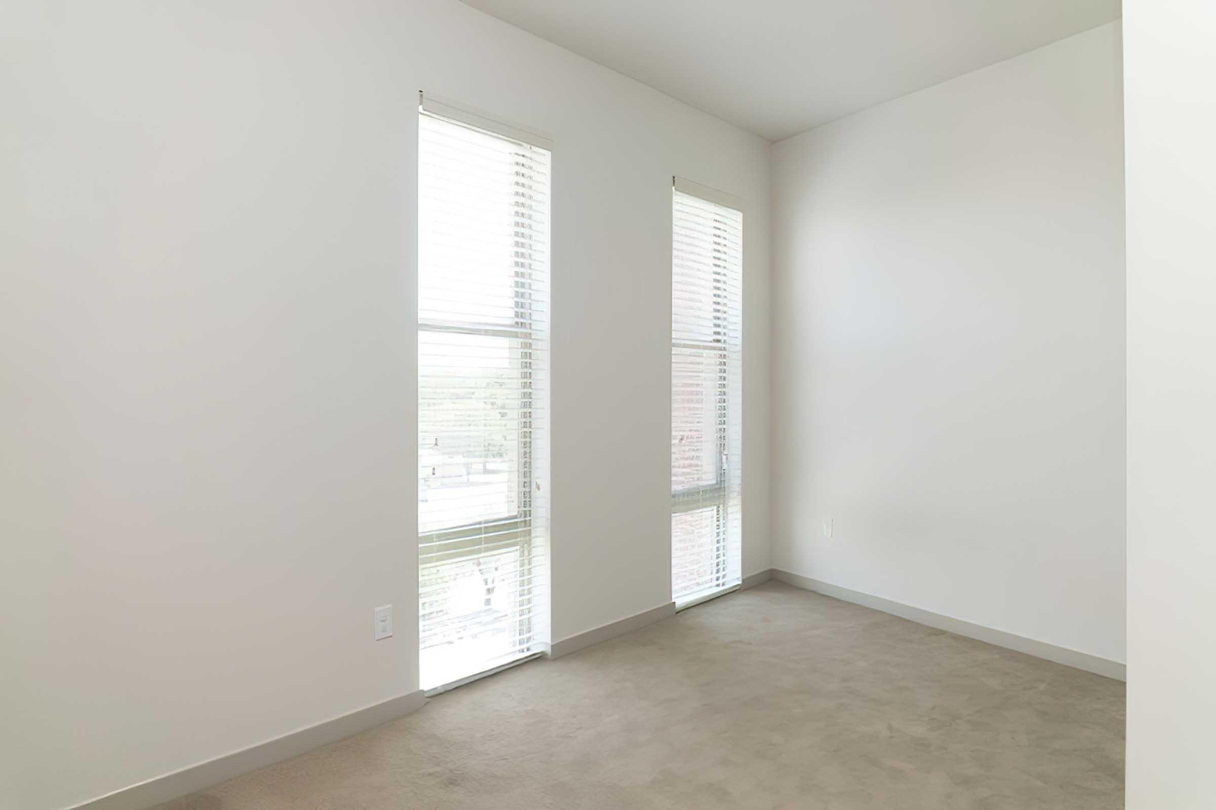 A spacious room with light-colored walls and beige carpet. It features two large windows with white blinds allowing natural light to enter.