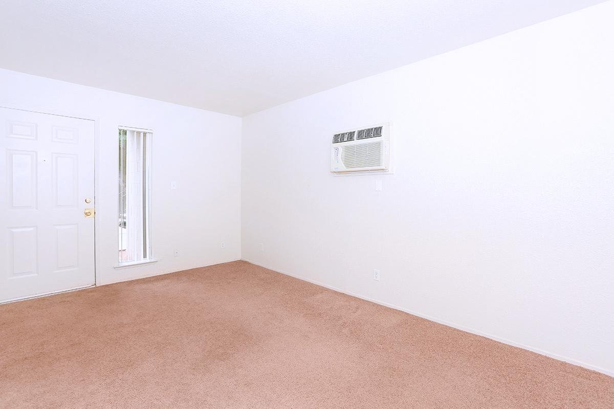 Spacious, empty room with light beige carpet. There is a front door on the left and a vertical window next to it. An air conditioning unit is mounted on the wall. The walls are painted white, creating a bright and open feel.