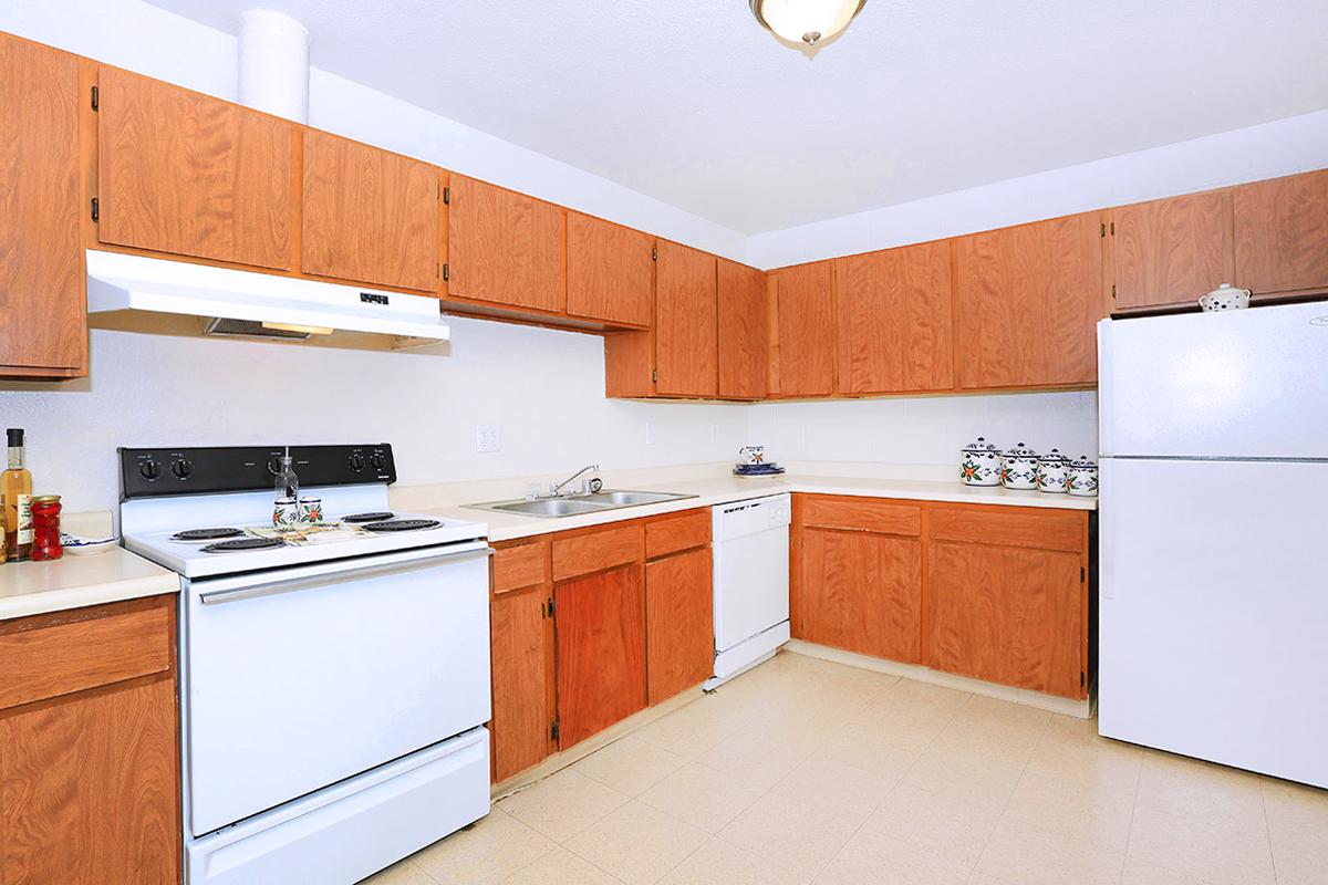 A tidy kitchen featuring wooden cabinets, a white stove, and a sink with a countertop. There’s a dishwasher next to the sink, and a white refrigerator stands against the wall. The space has a neutral-colored floor and is well-lit, creating a welcoming atmosphere.