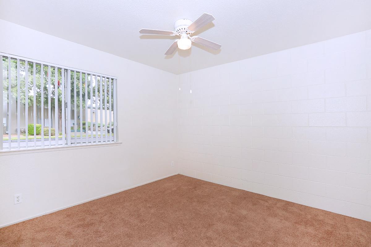 Empty room with light brown carpet, white walls, and a ceiling fan. A window with vertical blinds allows natural light to enter, revealing greenery outside. The room has a simple and spacious layout, suitable for various arrangements.