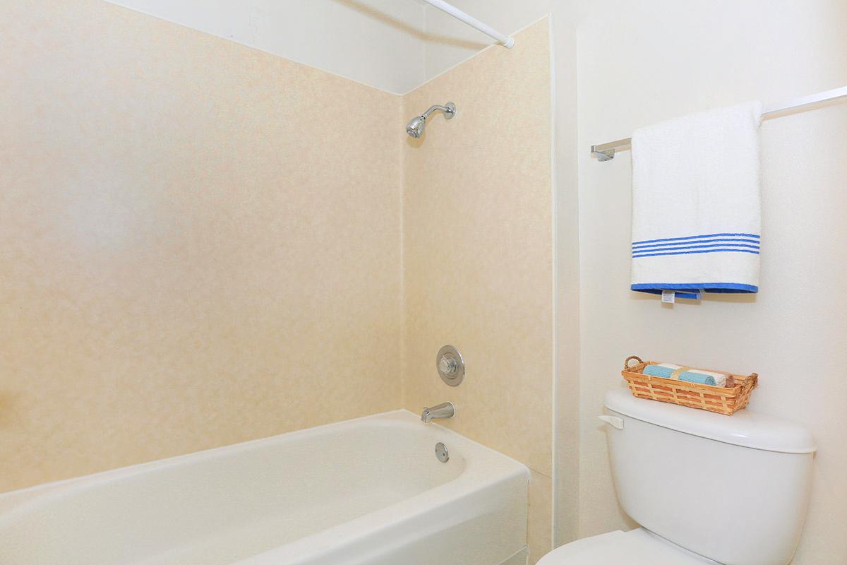 A clean bathroom featuring a white bathtub, a showerhead, and a beige wall. A white towel with blue stripes hangs on the towel rack, and a small basket with toiletries sits on top of the white toilet. The overall atmosphere is bright and simple.