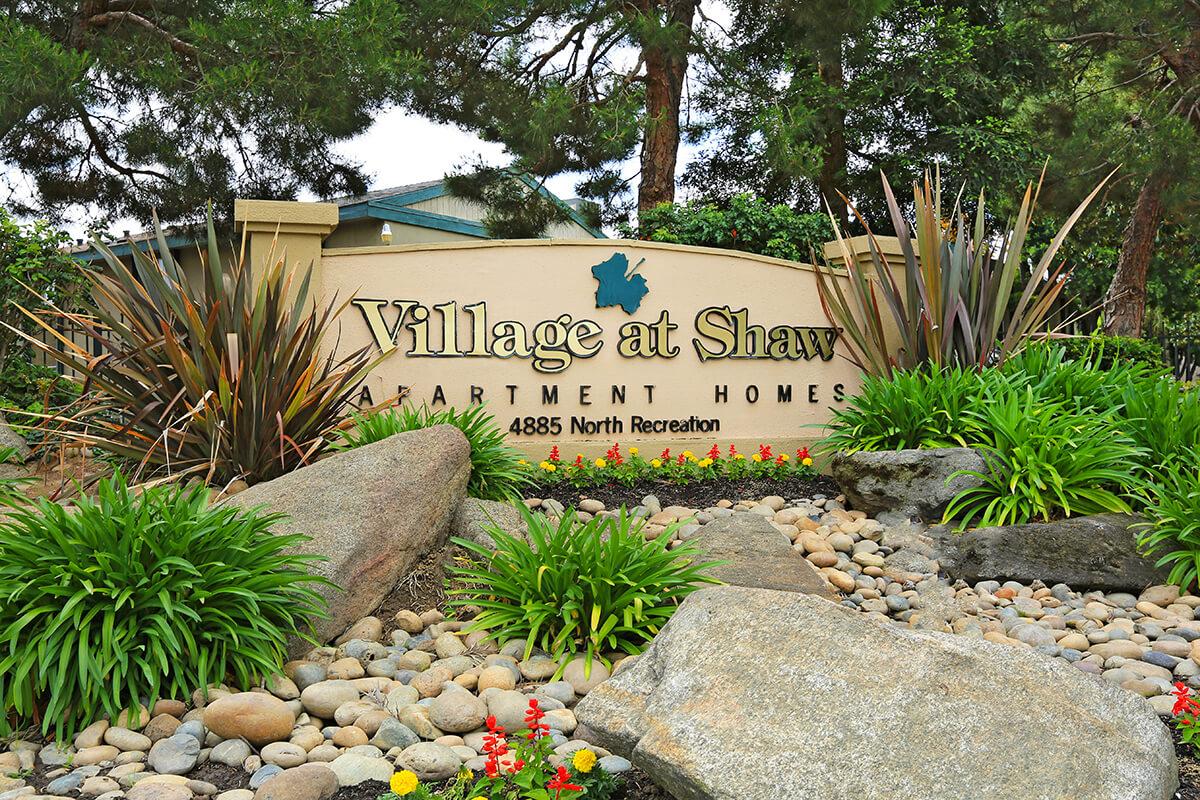 Sign for "Village at Shaw Apartment Homes" with the address "4885 North Recreation." The sign is surrounded by decorative landscaping, including rocks and colorful plants. The background features trees and a building structure.