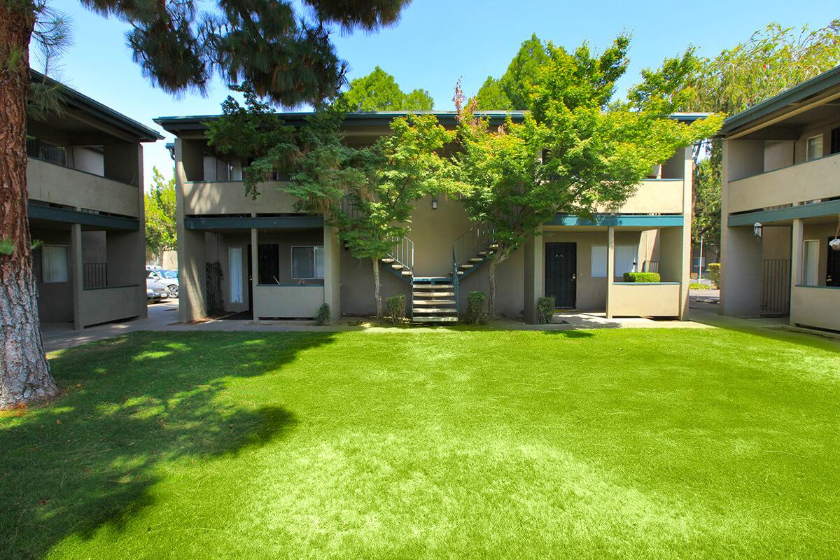 A well-maintained residential area featuring two-story apartment buildings surrounded by green trees. In the foreground, there is a vibrant, grassy lawn, and a set of stairs leads up to the apartments. The sky is clear and blue, creating a peaceful and inviting atmosphere.