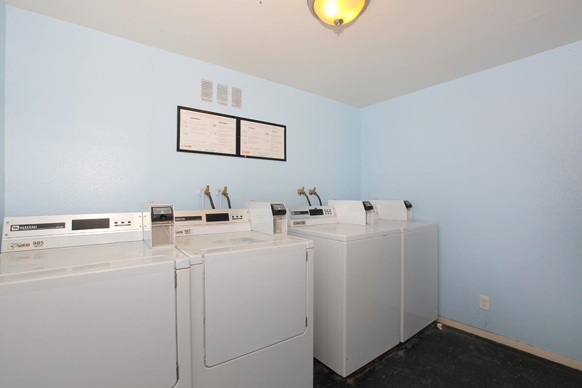 A bright laundry room featuring three white washing machines lined up against a light blue wall. Two framed certificates are mounted on the wall above the machines, and there is a ceiling light fixture for illumination. The floor is dark and plain, creating a simple, functional space.