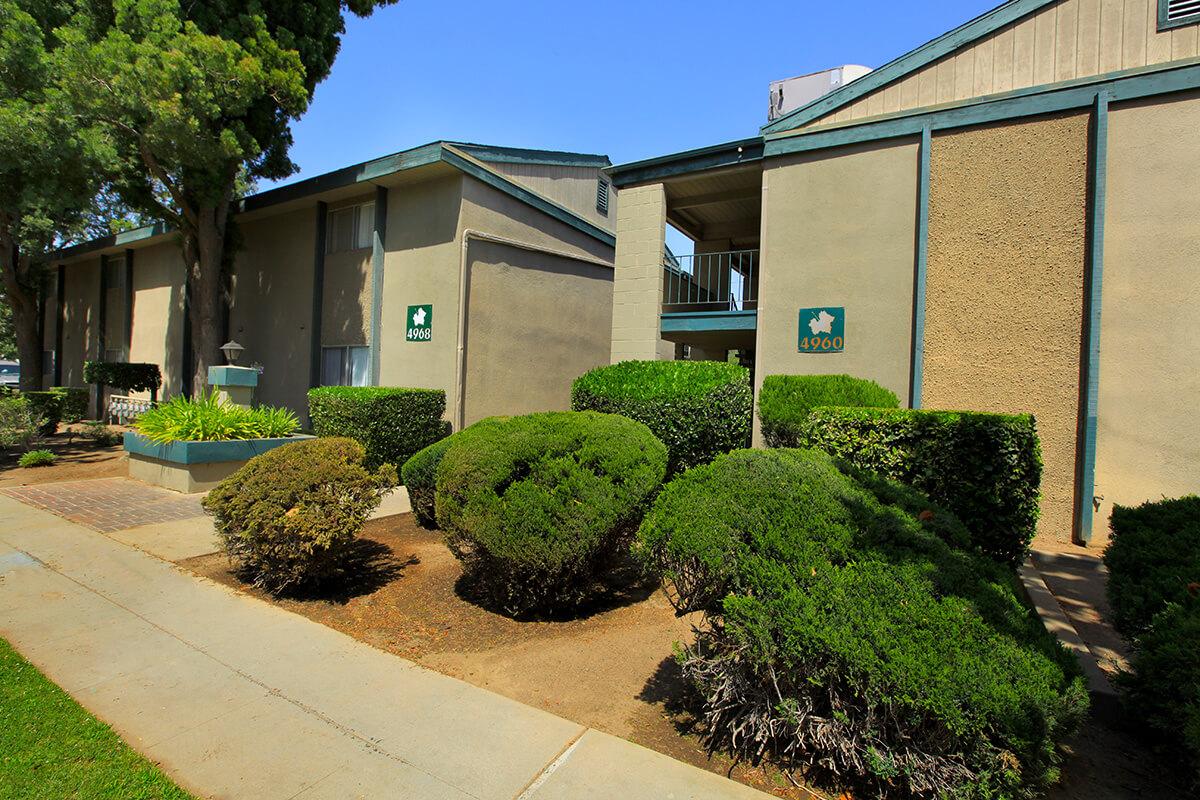 A view of a residential building, featuring two stories with green accents. Bushy shrubs and neatly trimmed plants line the walkway, which leads to the entrance. The building has signs displaying the numbers "4900" and "4902." A tree provides shade to the scene, enhancing the greenery.