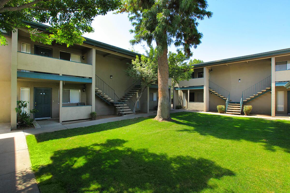 A view of a two-story apartment complex featuring an open courtyard with grassy areas and trees. The building has staircases leading to the upper level apartments, and there are potted plants near the entrance of the units. Bright blue sky and sunlight enhance the inviting atmosphere.