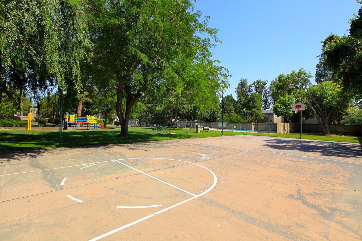 A sunny park featuring a basketball court in the foreground, surrounded by lush green trees. In the background, there is a colorful children's playground and open grassy areas. The sky is clear and blue, creating a bright and cheerful atmosphere.
